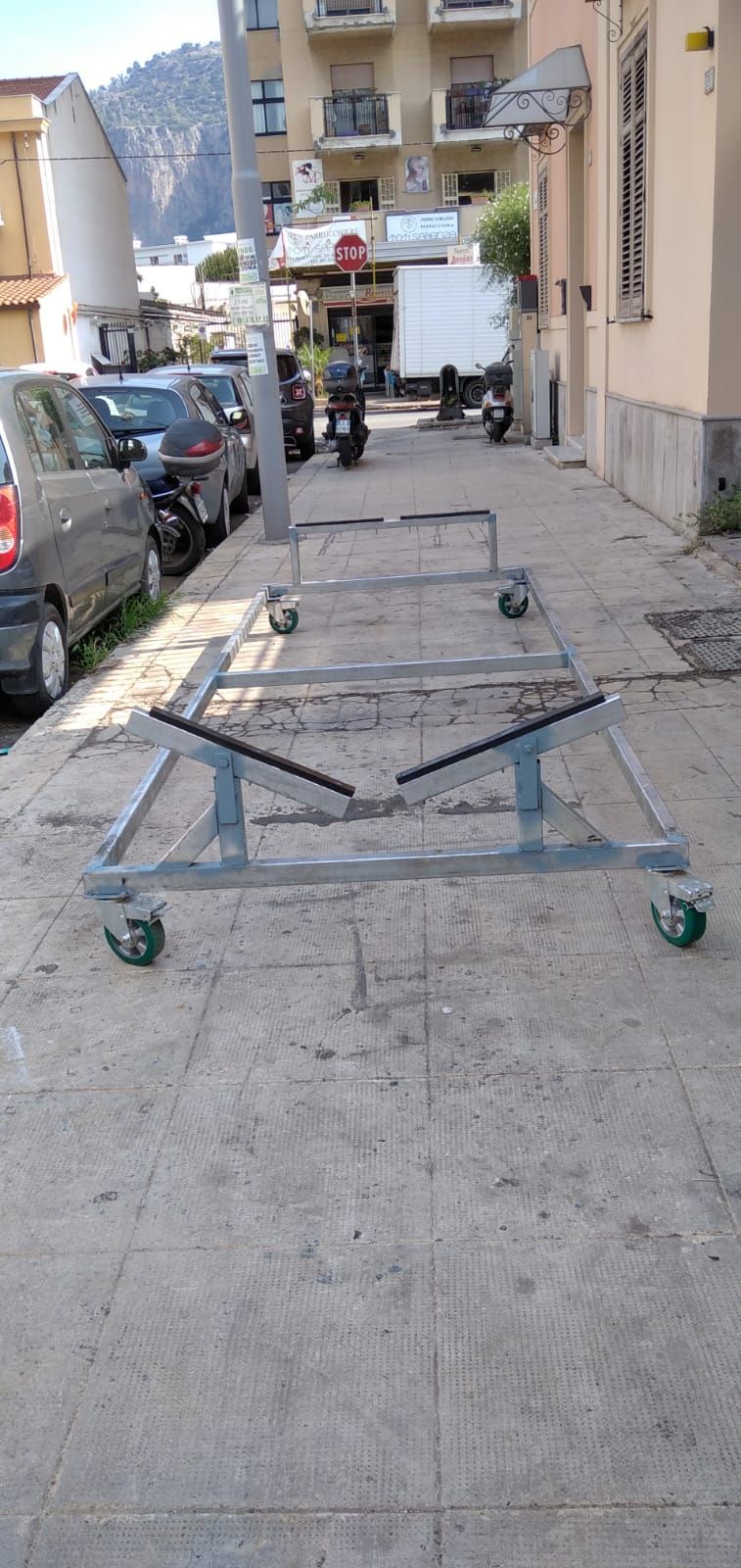 Un carrello metallico su ruote è parcheggiato su un marciapiede, accanto ad auto parcheggiate ed edifici.