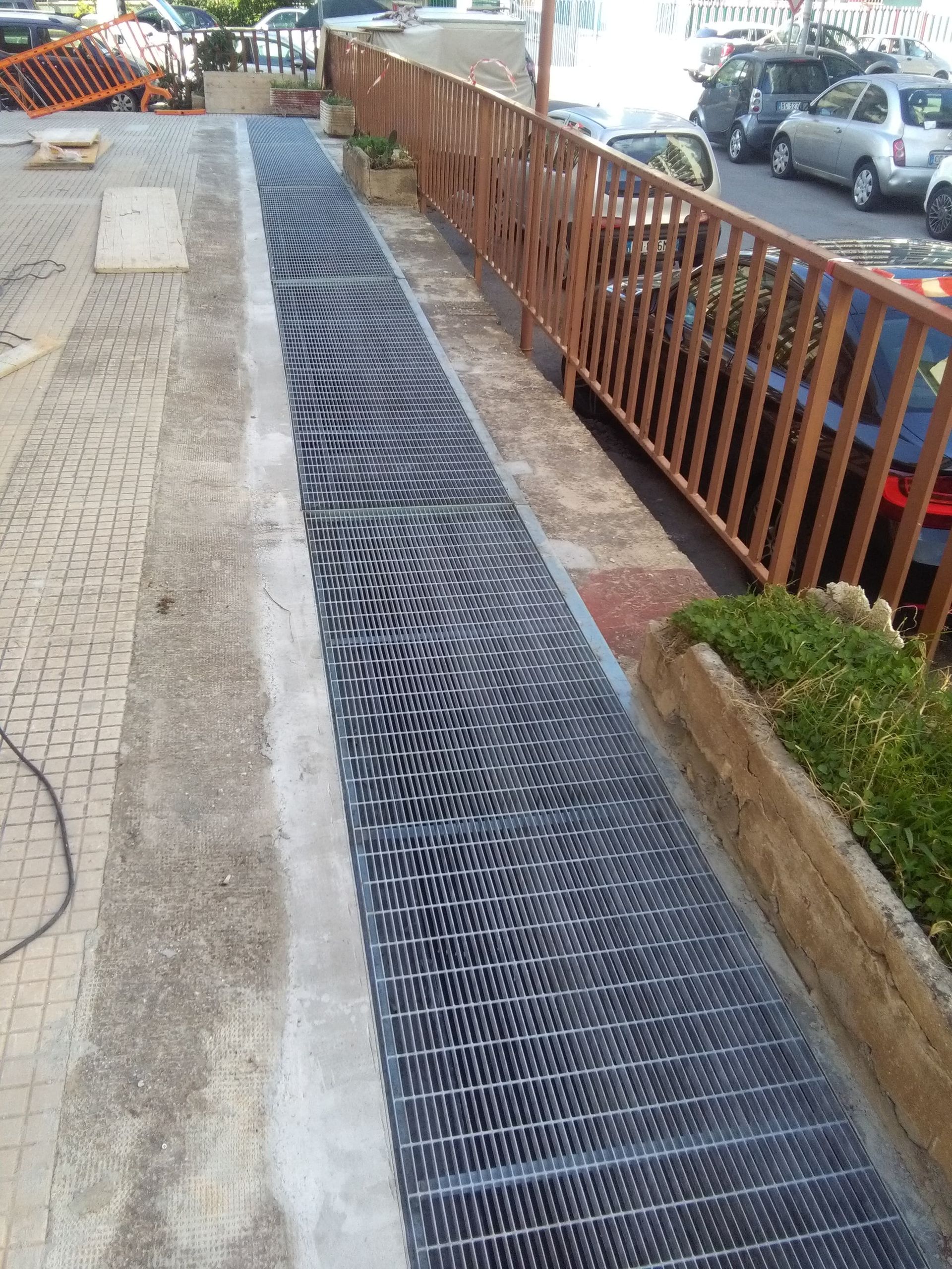 Sistema di drenaggio in griglia metallica lungo un marciapiede. Ringhiera in legno e auto parcheggiate sullo sfondo.