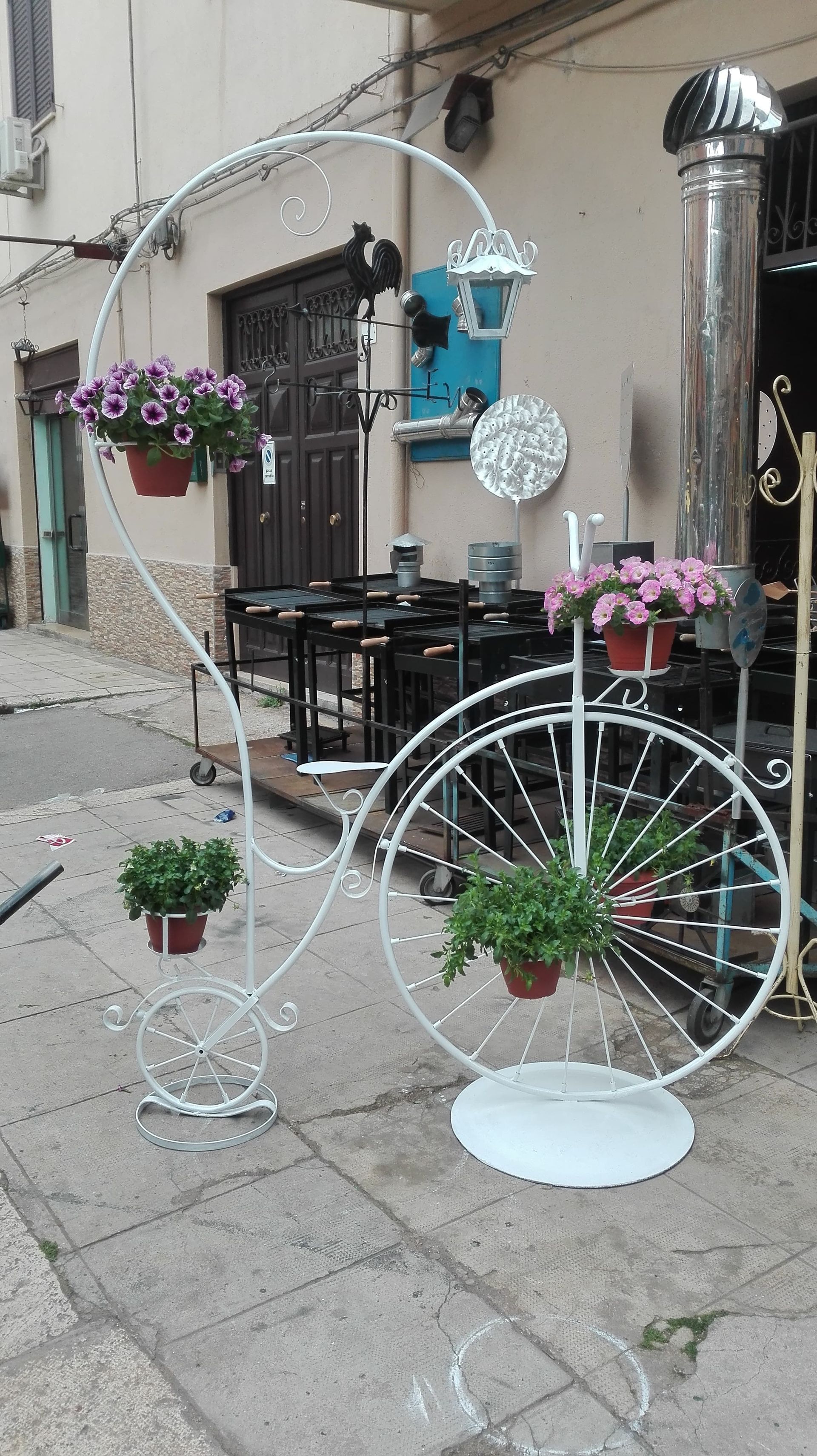 Supporto per piante in metallo bianco con ruota di bicicletta attaccata, che sostiene fiori in vaso su un marciapiede.