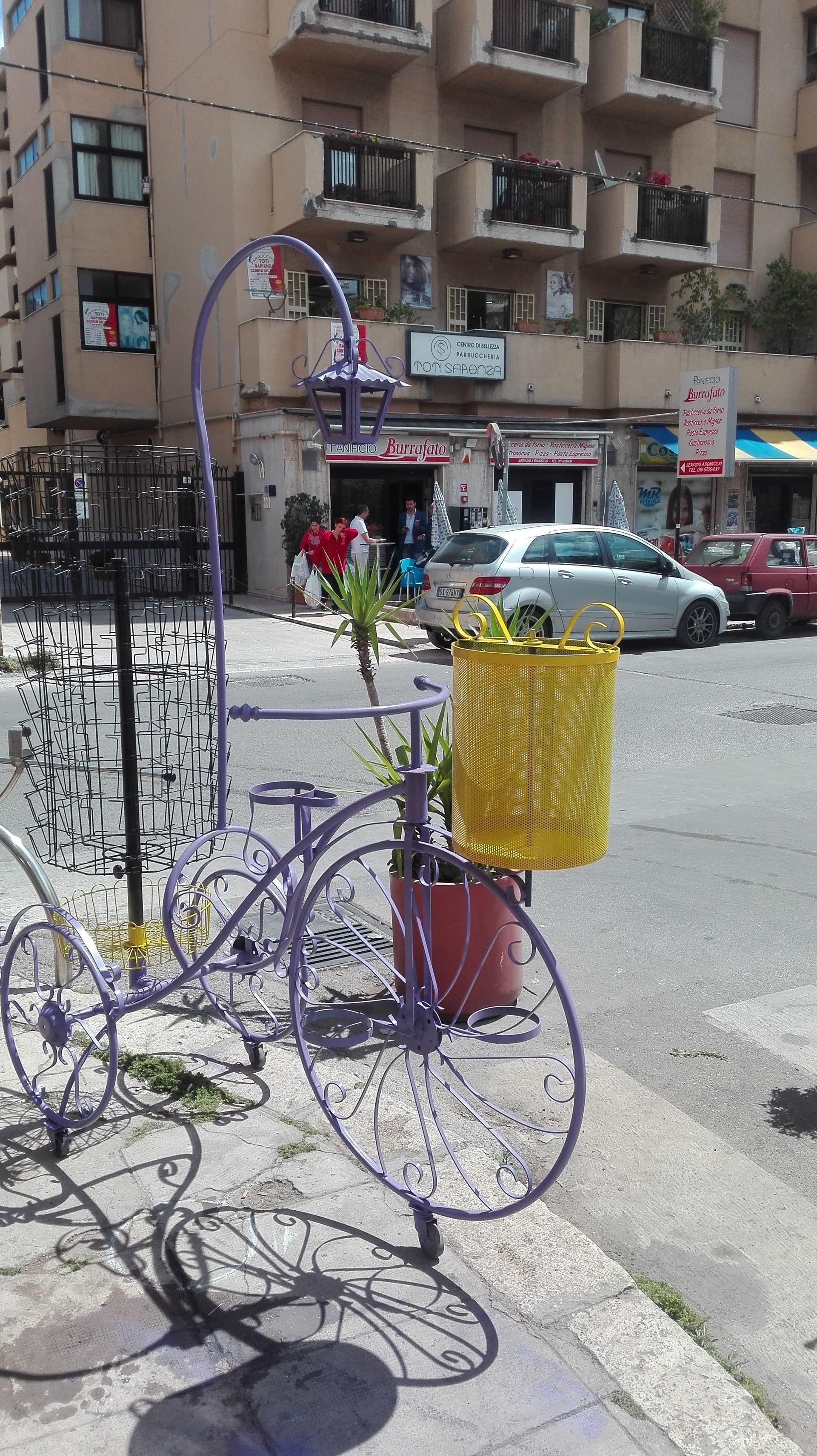 Scultura di bicicletta viola con cestino giallo, ambientazione stradale.