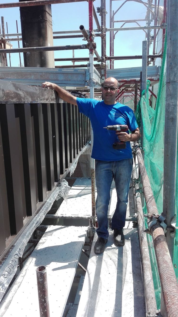 Uomo in camicia blu e jeans su un'impalcatura, con in mano un trapano, che indica una struttura scura.