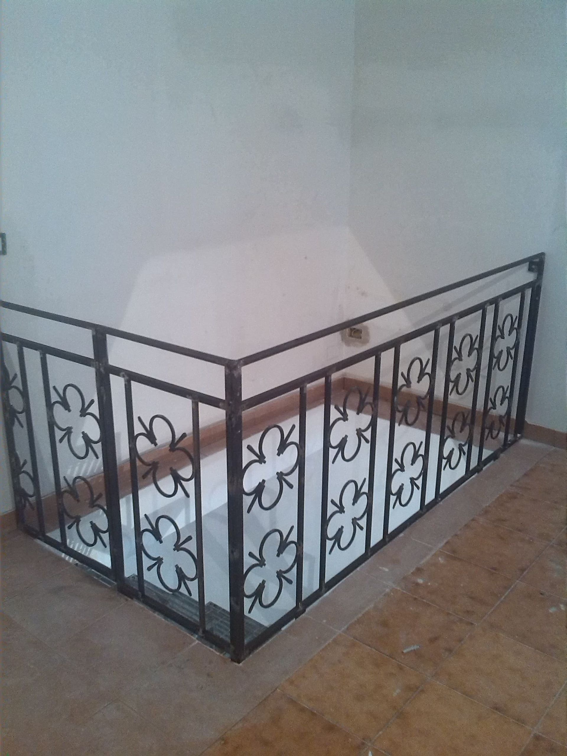 Ringhiera in ferro battuto nero attorno a una zona incassata, contro pareti chiare e pavimento in legno.