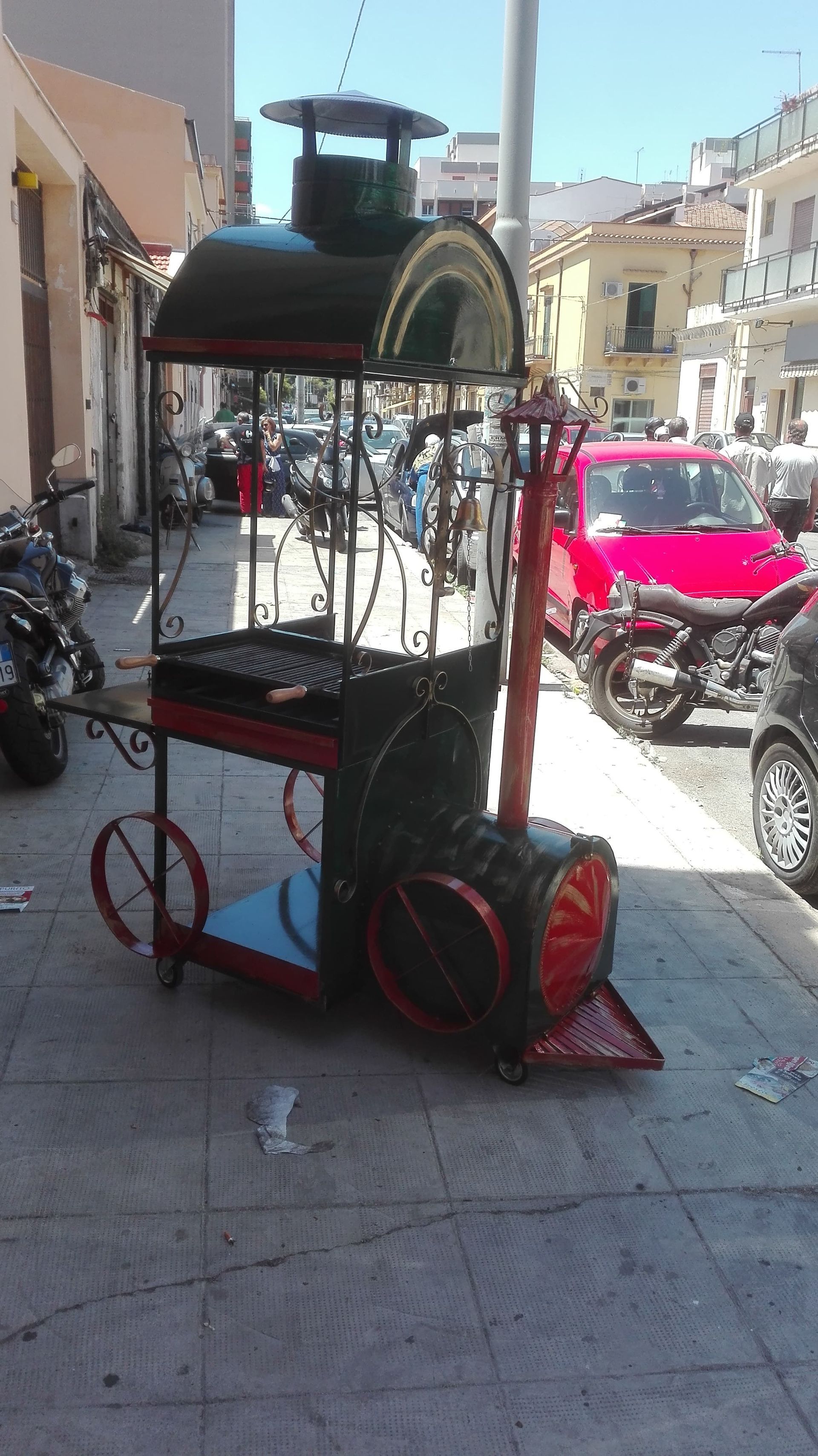 Griglia da esterno a forma di treno su un marciapiede. Metallo nero e rosso, con griglia. Motociclette ed edifici sullo sfondo.