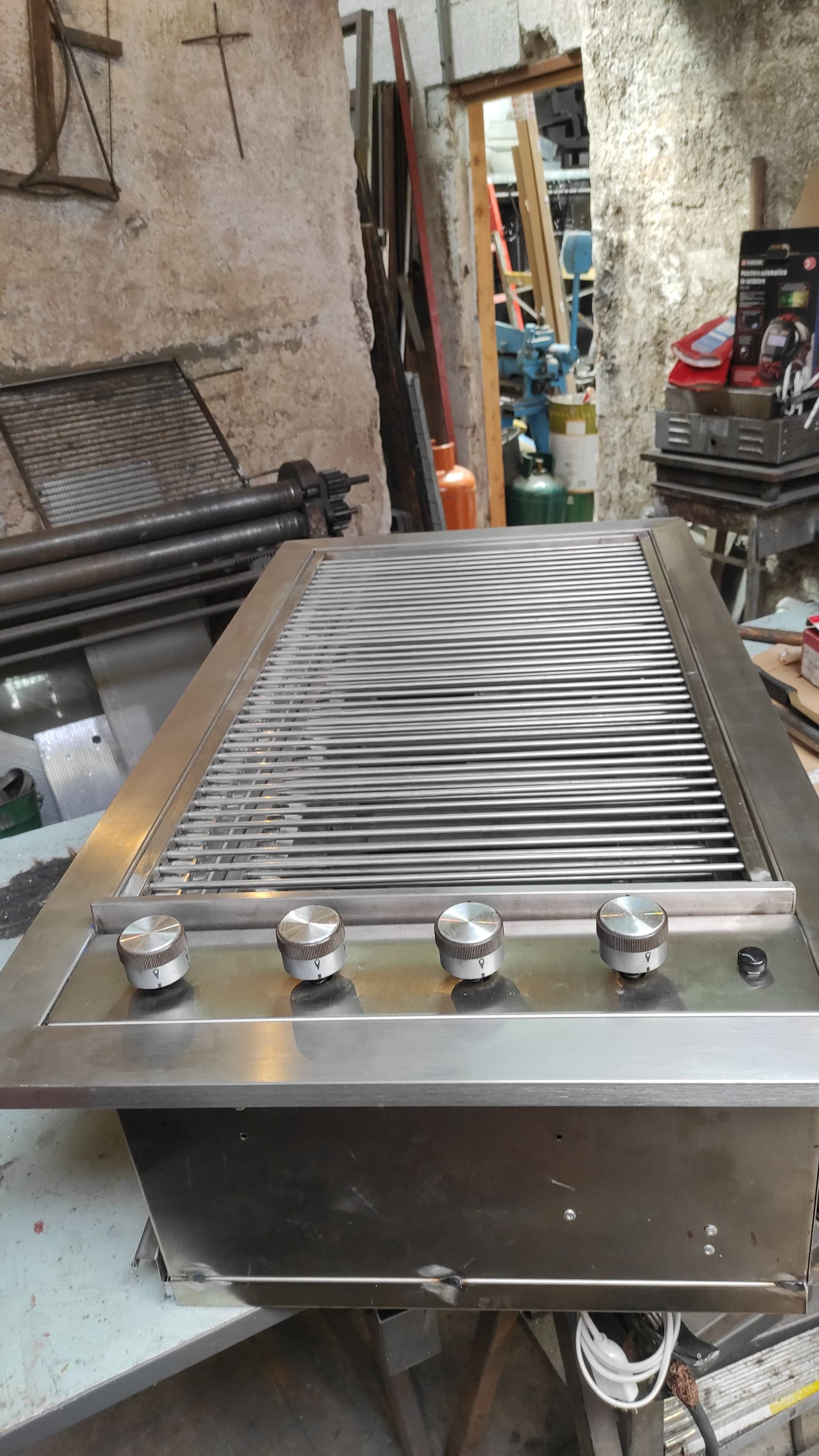 Griglia a gas in acciaio inox con quattro manopole, in un'officina disordinata.