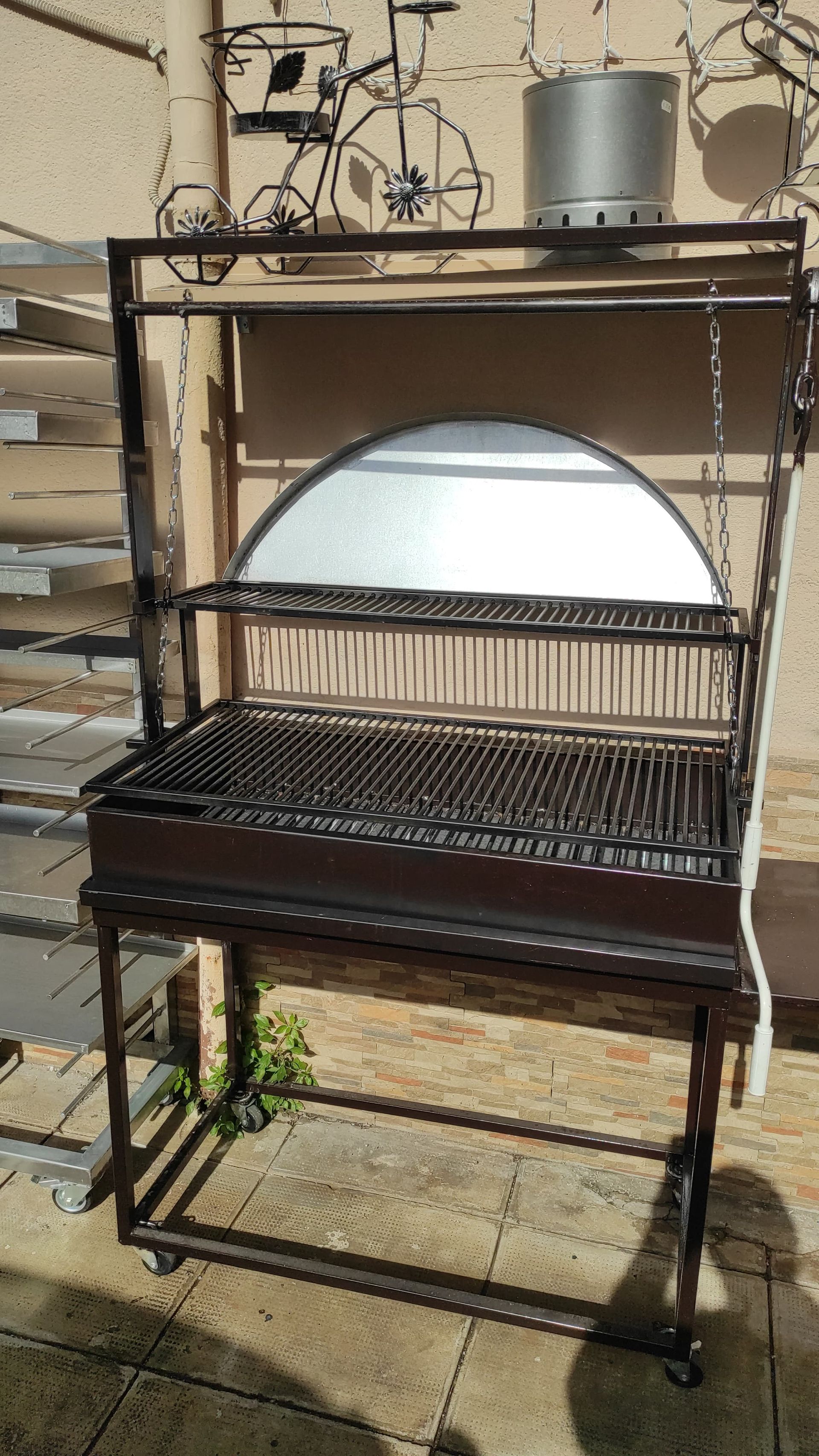 Griglia metallica con ripiani, ruote e coperchio metallico, posizionata all'esterno.