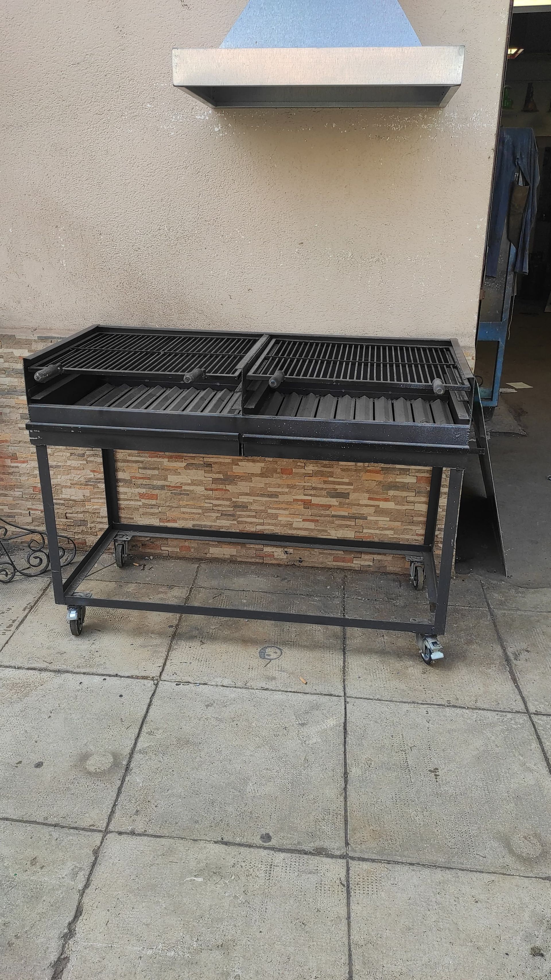 Griglia metallica nera su ruote all'aperto, vicino a un muro di pietra, con una presa d'aria superiore.