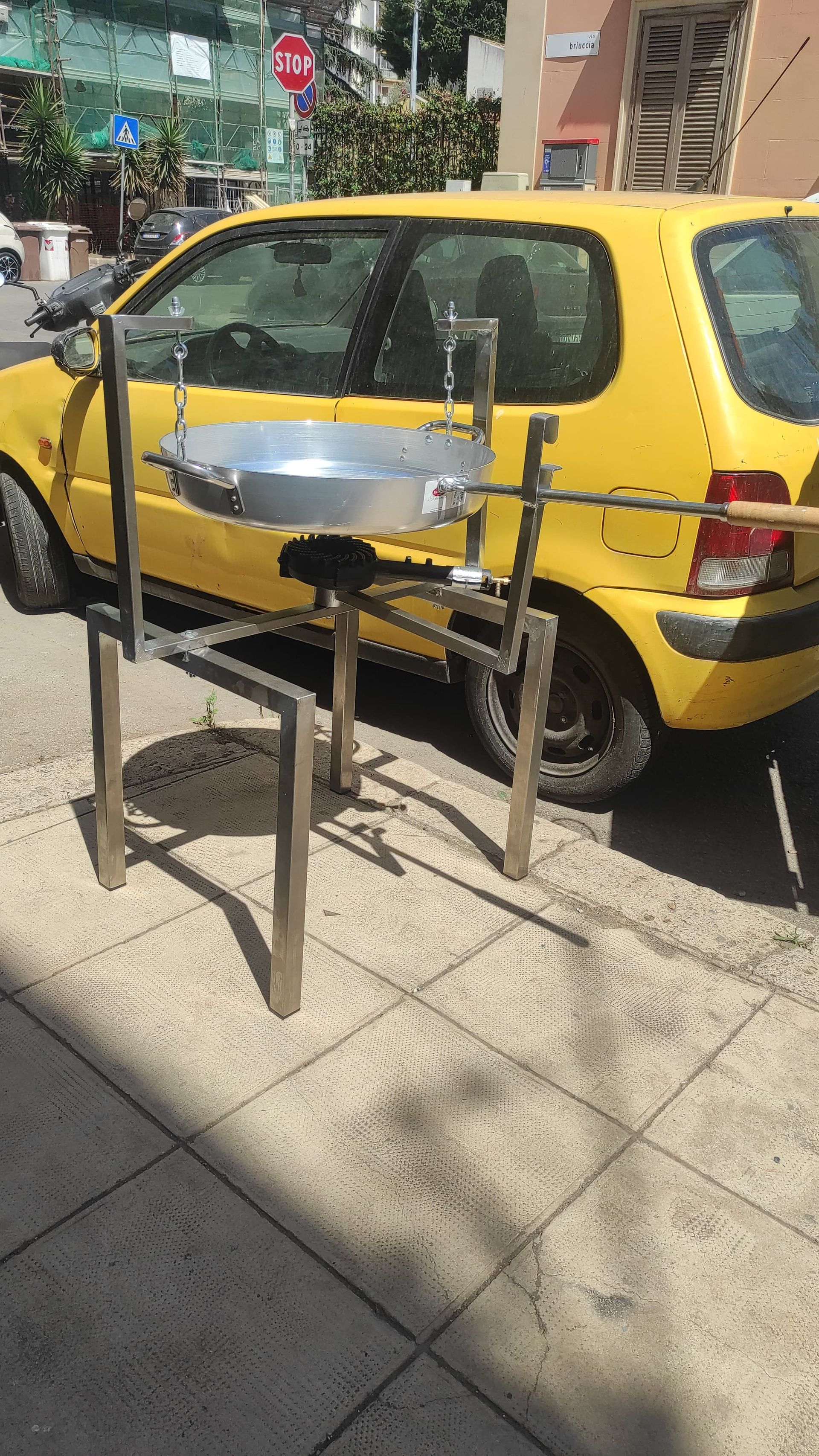 Un supporto di metallo con una padella d'argento sopra, accanto a un'auto gialla parcheggiata su un marciapiede.