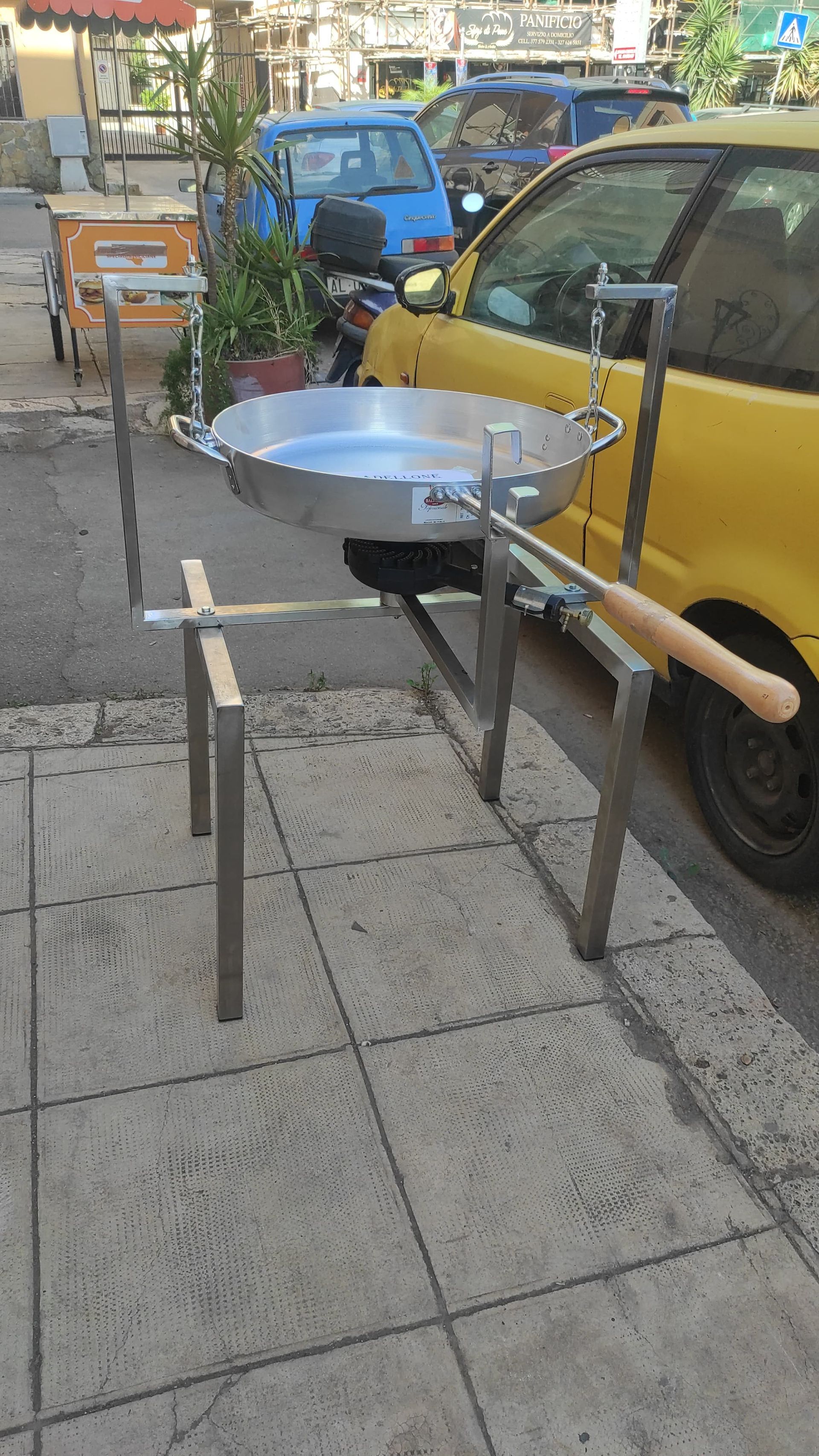 Una padella per paella in metallo su un supporto, all'aperto. Un'auto gialla è parcheggiata lì vicino.