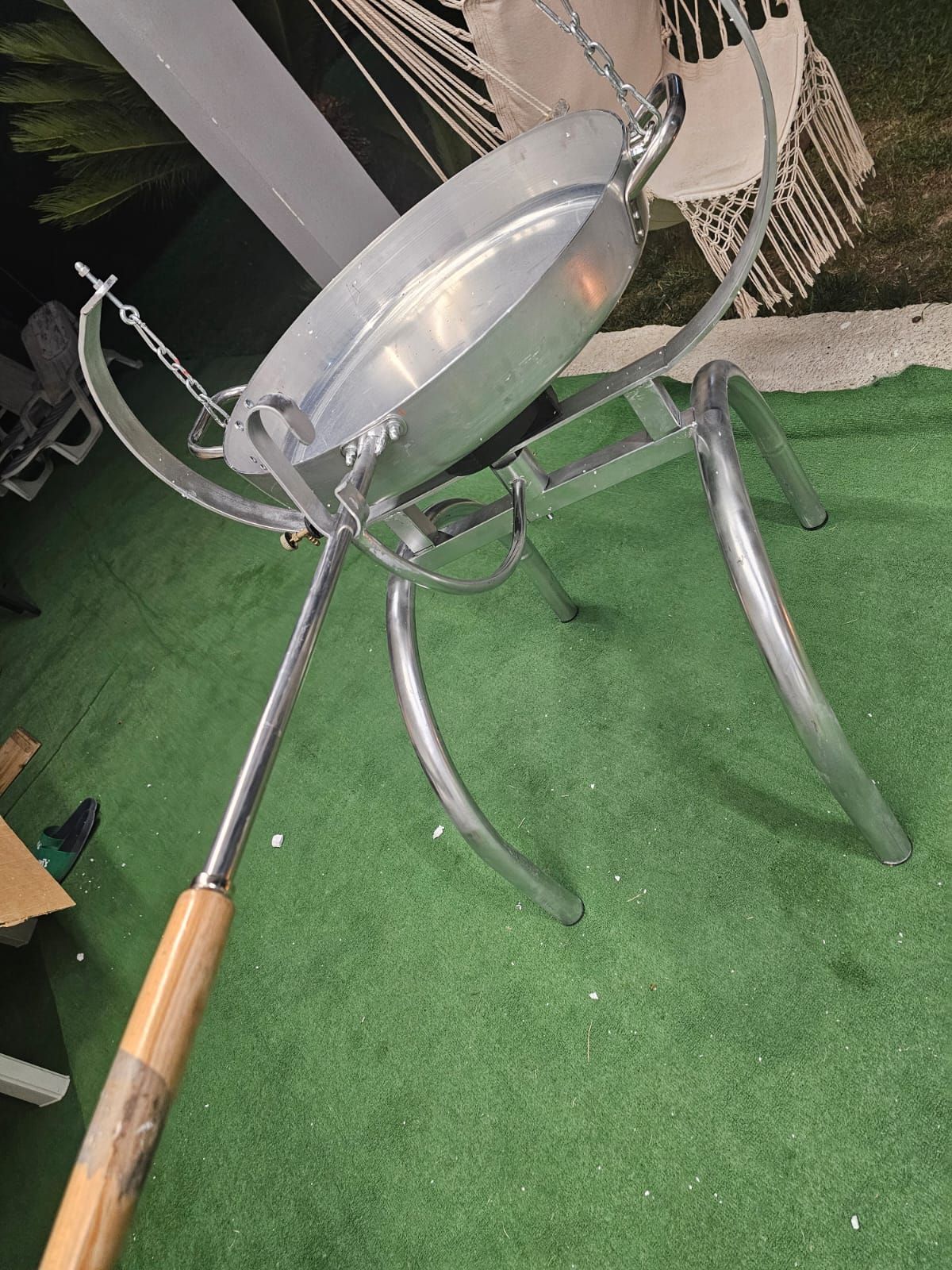 Pentola da cucina in metallo per esterni su treppiede con lungo manico in legno, appoggiata su erba verde.