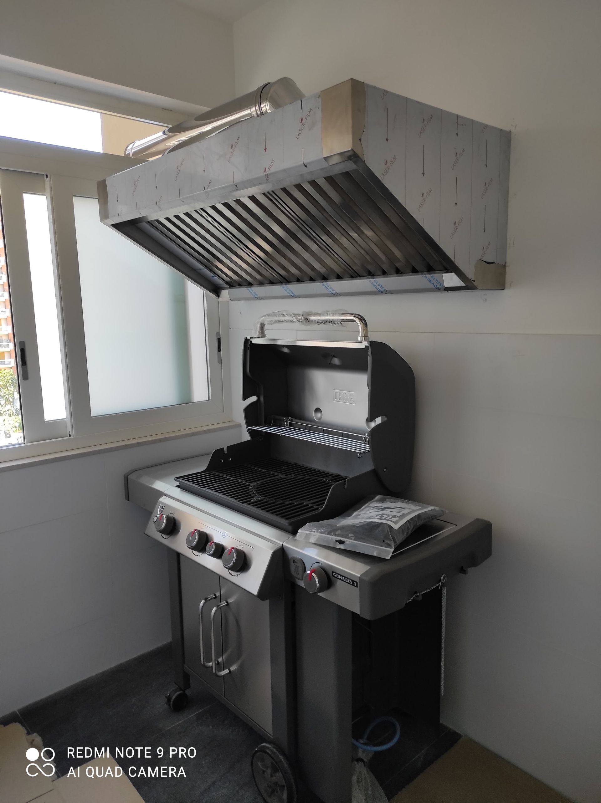 Griglia con cappa di ventilazione fissata a una parete, accanto a una finestra.