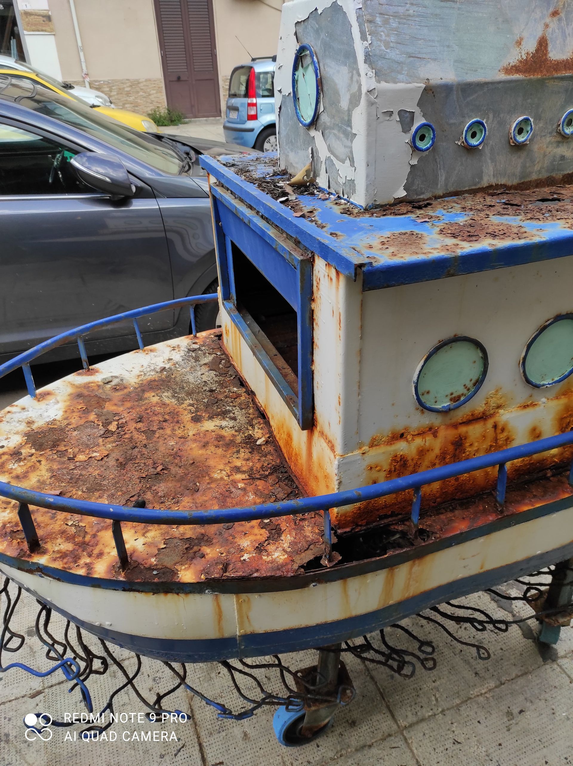 Barca giocattolo arrugginita su ruote, vernice bianca e blu, all'aperto.