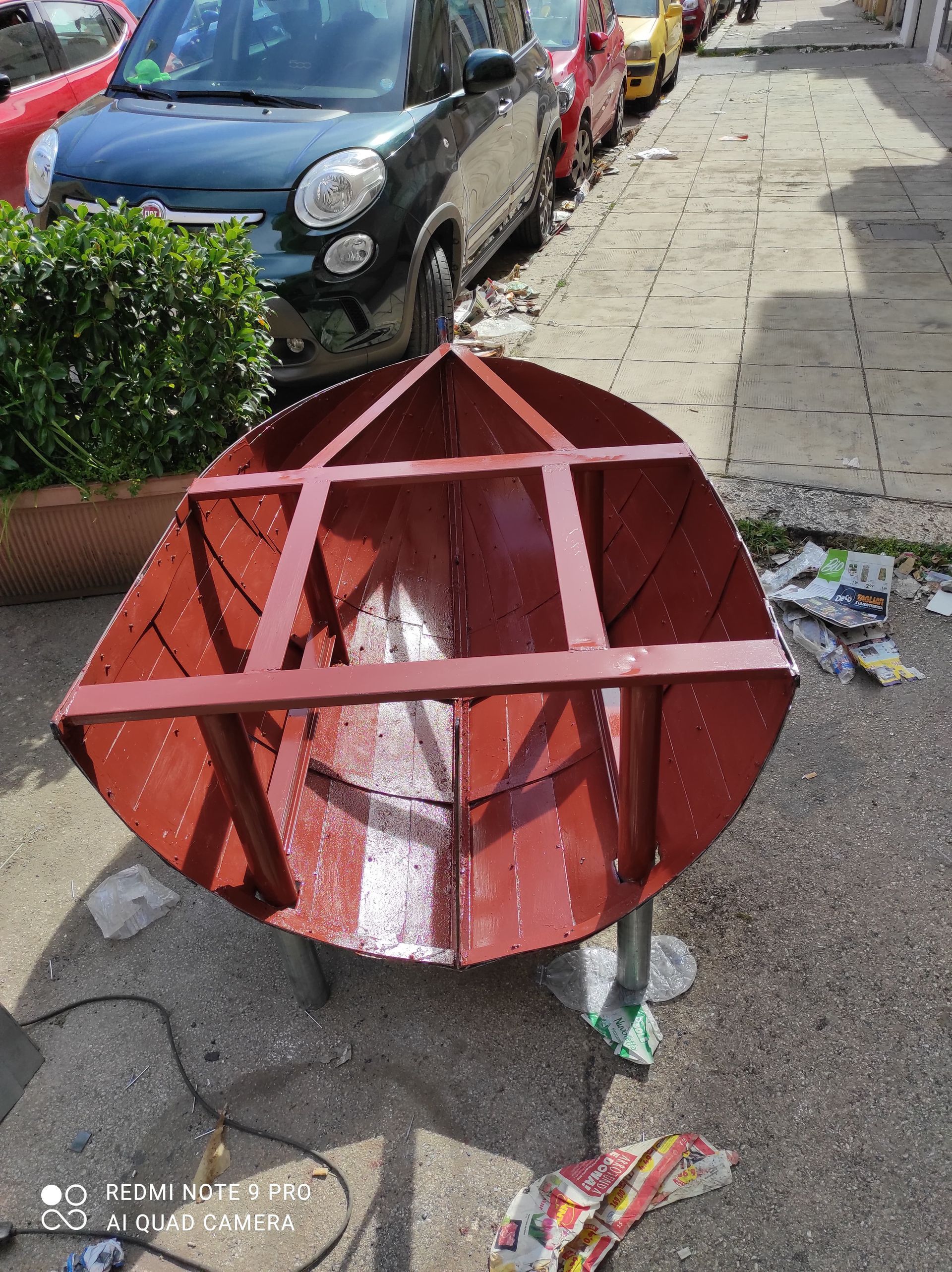 Telaio di una barca in metallo rosso su un marciapiede; auto e detriti sullo sfondo.