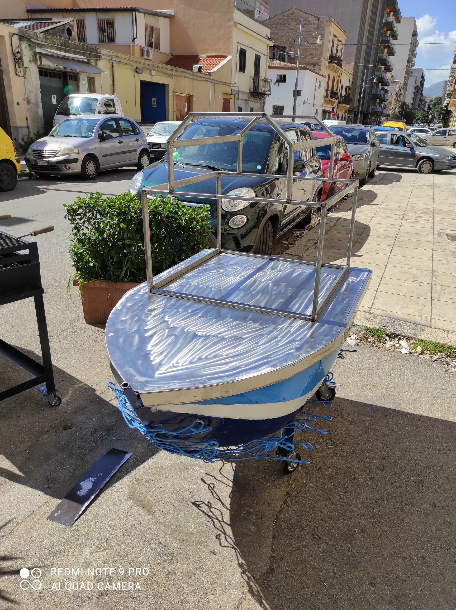 Una piccola barca blu e bianca su ruote, avvolta nella plastica, è parcheggiata su un marciapiede accanto a una strada con auto parcheggiate.