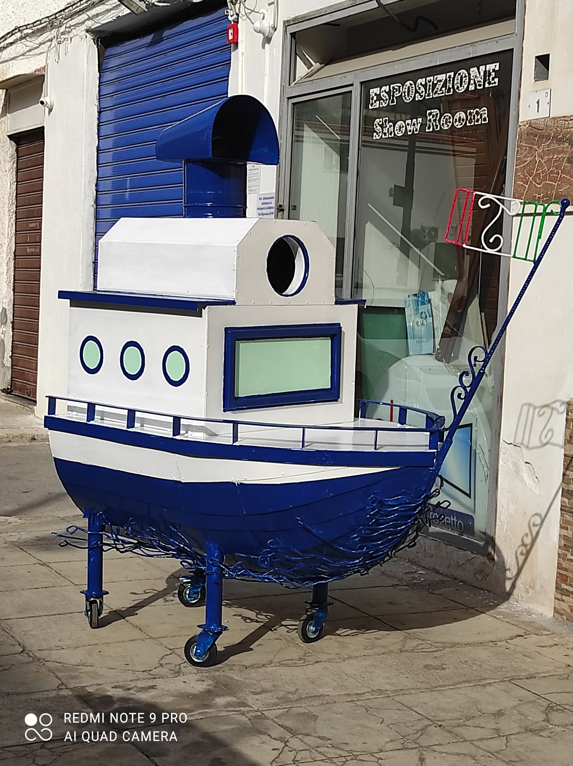 Carretto blu e bianco a forma di barca con ruote parcheggiato fuori da un negozio.