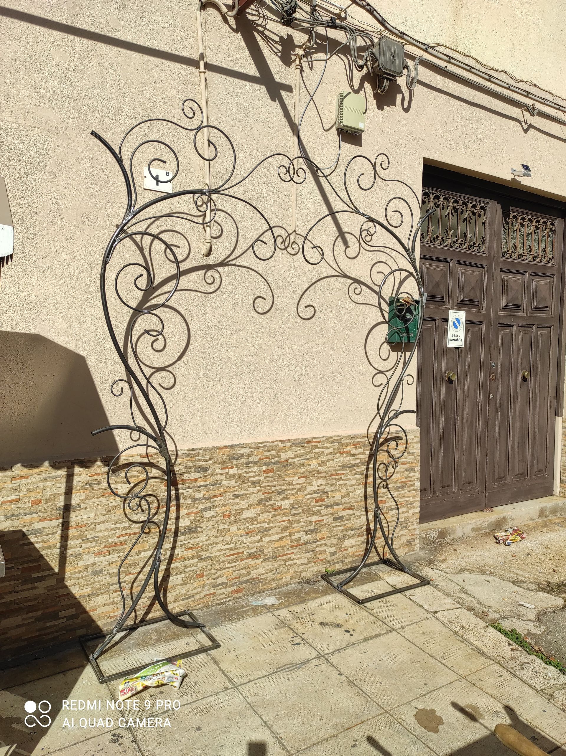 Arco in ferro battuto nero con vortici decorativi, appoggiato su un marciapiede contro un muro marrone chiaro e una porta marrone.