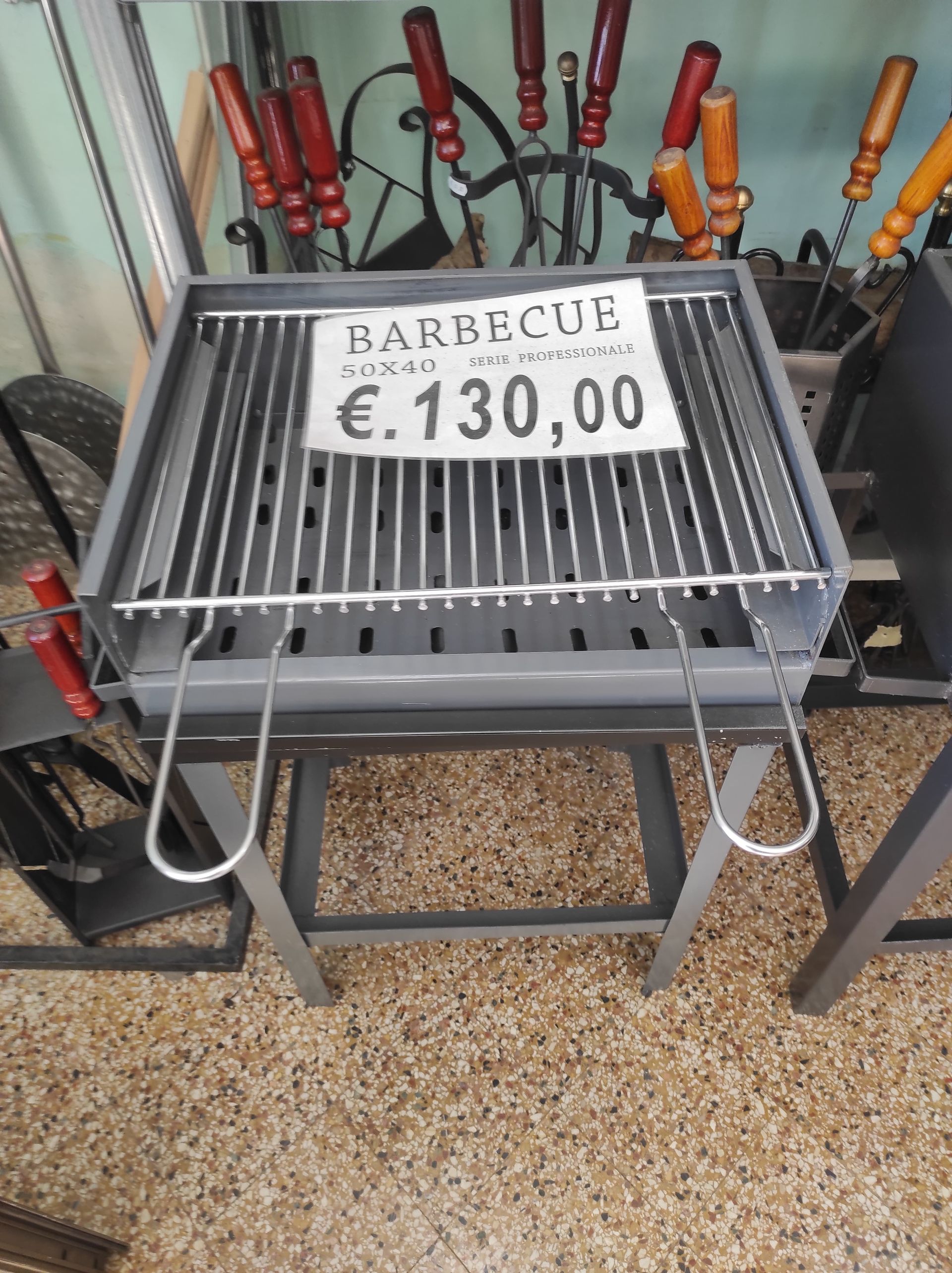 Griglia per barbecue in metallo grigio su supporto, al prezzo di € 130,00, con utensili per grigliare sullo sfondo.