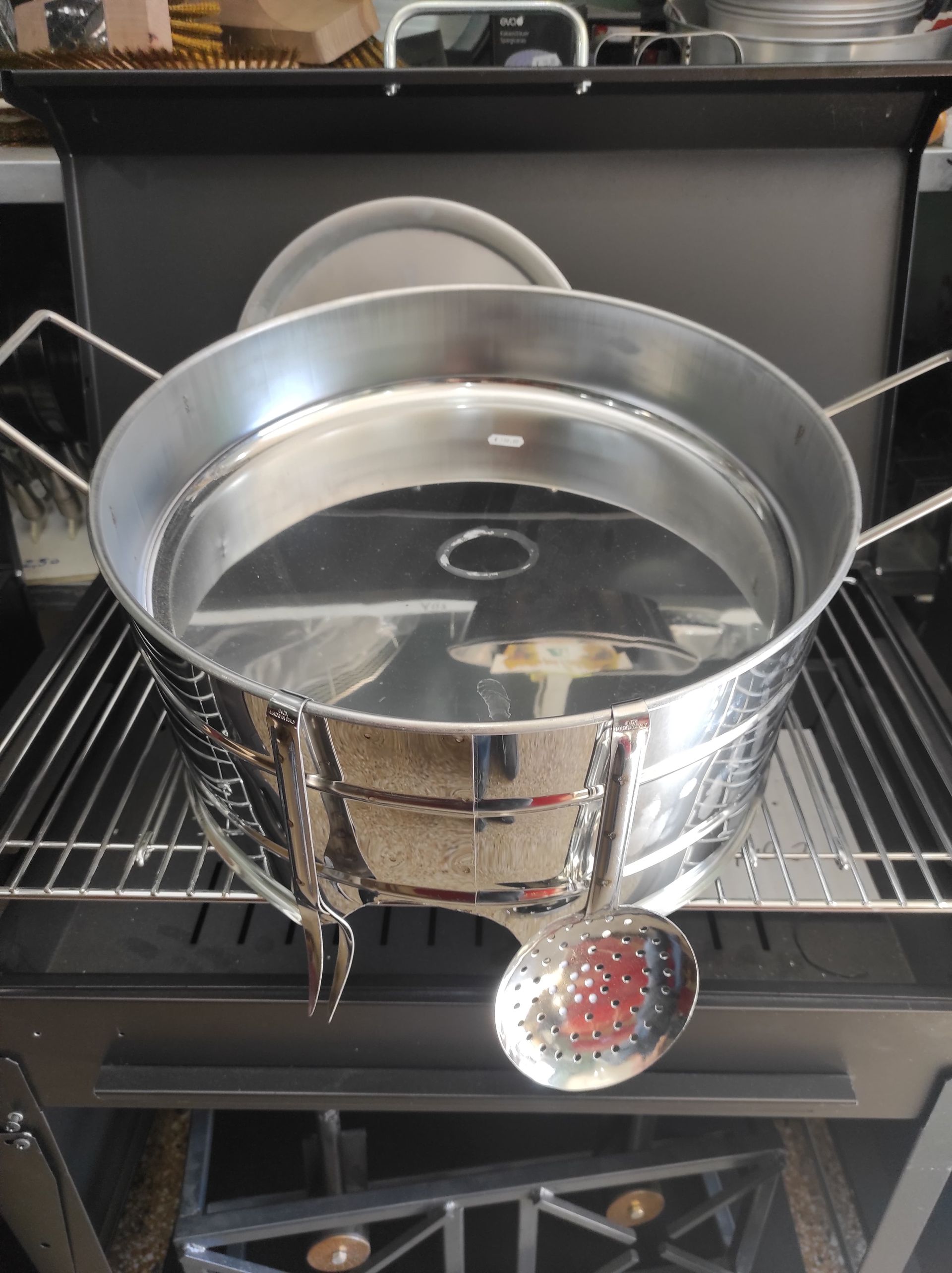 Pentola di metallo lucido su una griglia, con un colino attaccato. La pentola ha i manici ed è posizionata all'aperto.