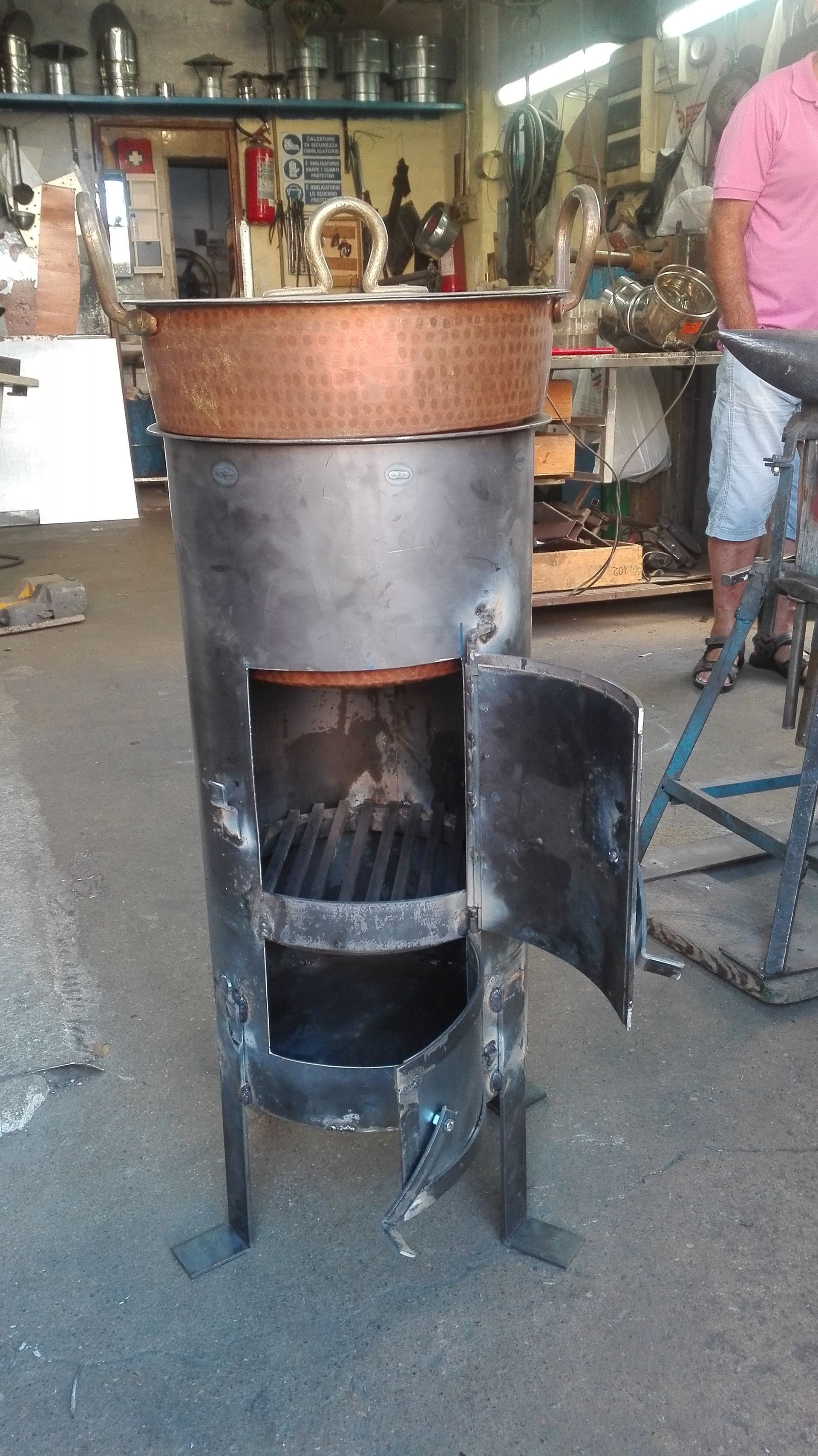 Una stufa di metallo con lo sportello aperto e il piano superiore in rame, in un'officina. Una persona è in piedi nelle vicinanze.
