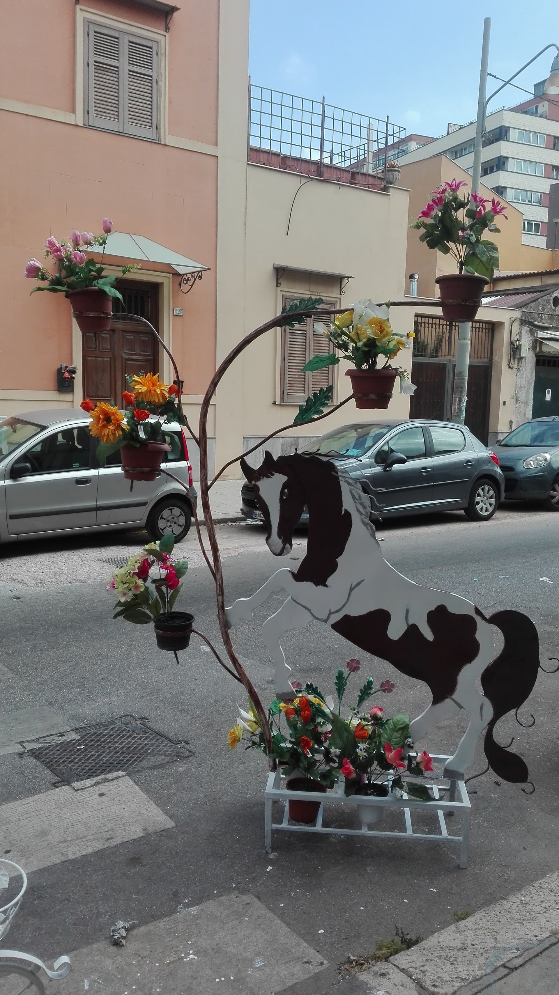 Decorazione stradale con un cavallo di metallo e vasi di fiori; sullo sfondo automobili ed edifici.
