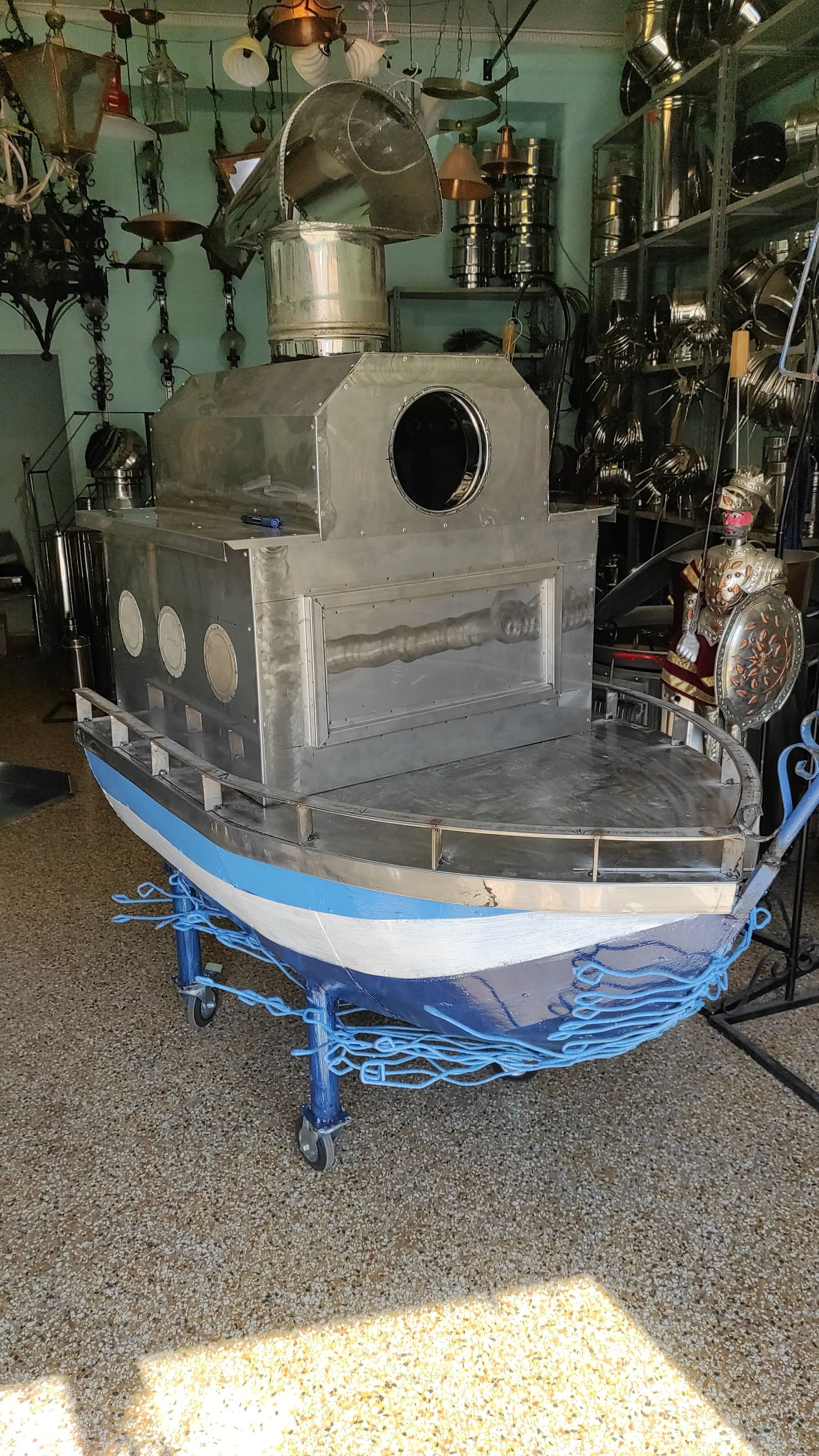 Forno a legna in metallo a forma di barca, scafo blu e bianco, su carrello con ruote blu, con camino.