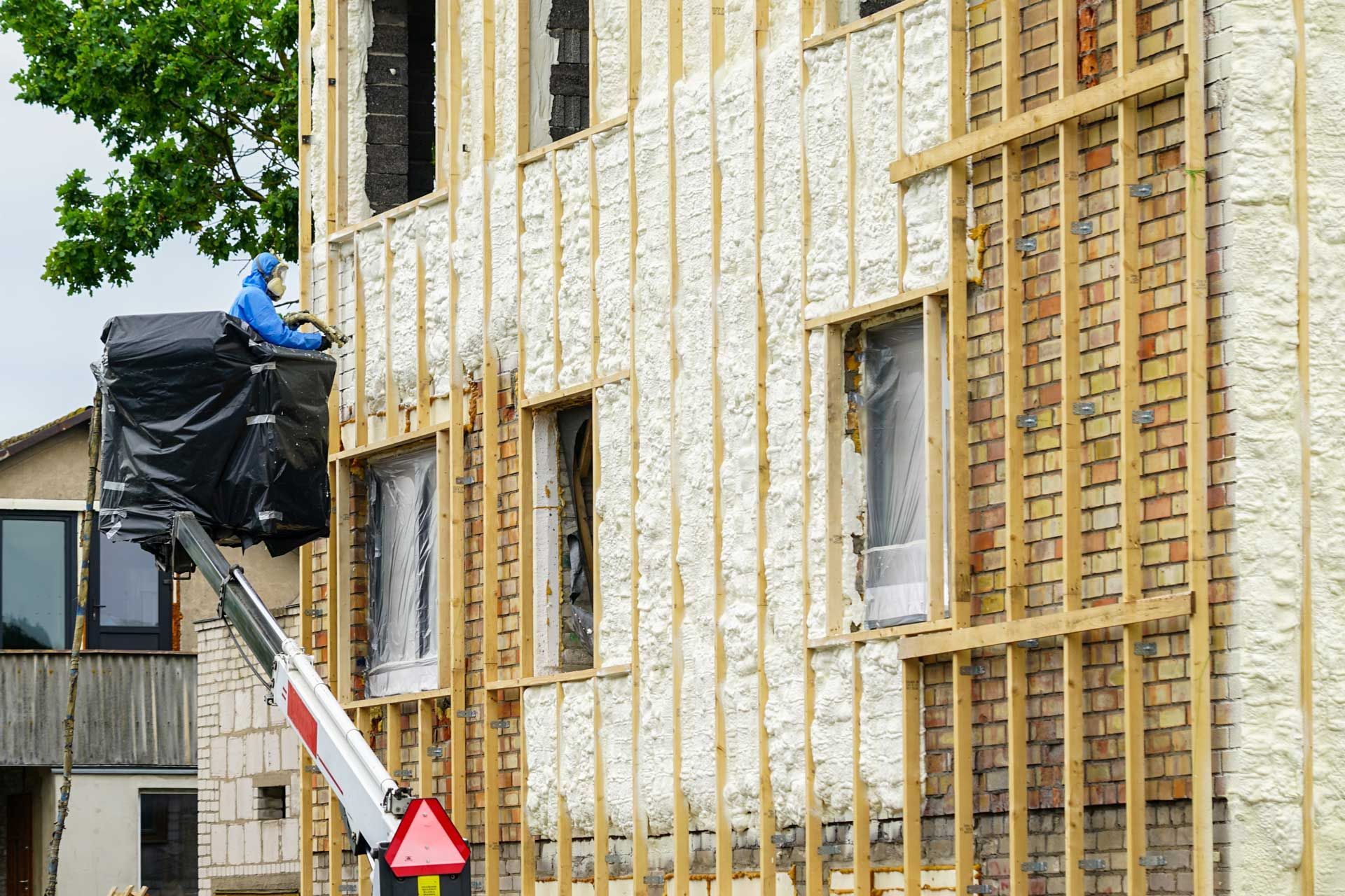 Man in blue protective gear sprays foam insulation on a building facade from a lift.