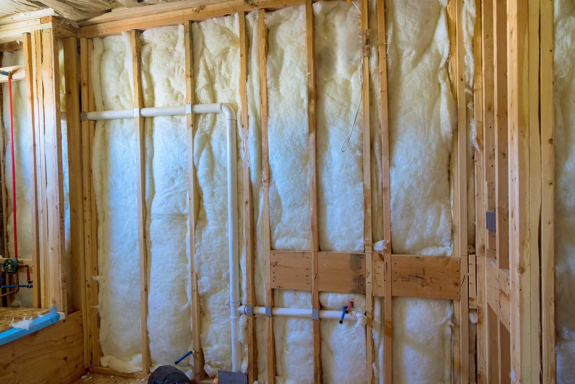 Interior wall under construction with insulation between wooden studs, showing pipes.
