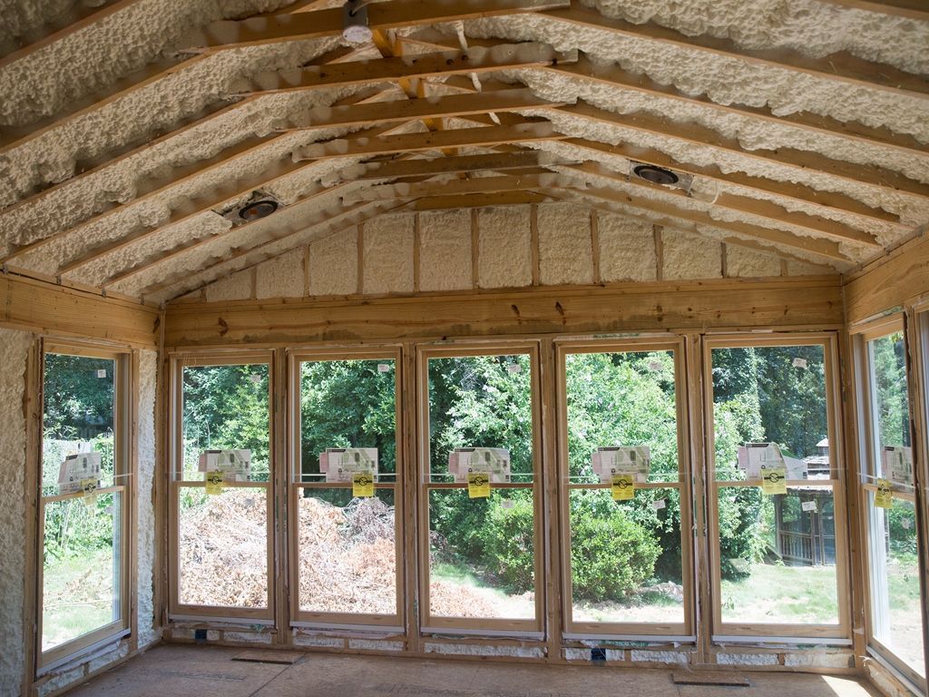 Interior of sunroom under construction, insulated with spray foam, windows overlooking greenery.