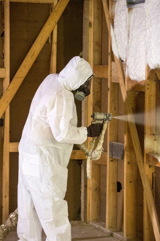 Person in white protective suit sprays insulation foam in a wooden wall framework.