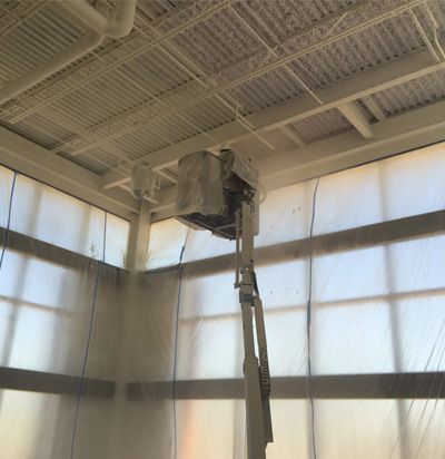 A white lift truck spraying white paint on the ceiling of a room, with plastic sheeting covering the windows.