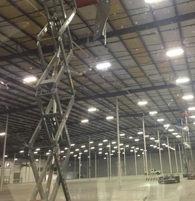 Scissor lift inside a large warehouse, reaching towards the ceiling; many lights and supports.