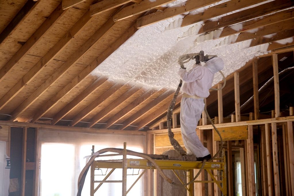Man in protective suit spraying foam insulation into a wood-framed ceiling.