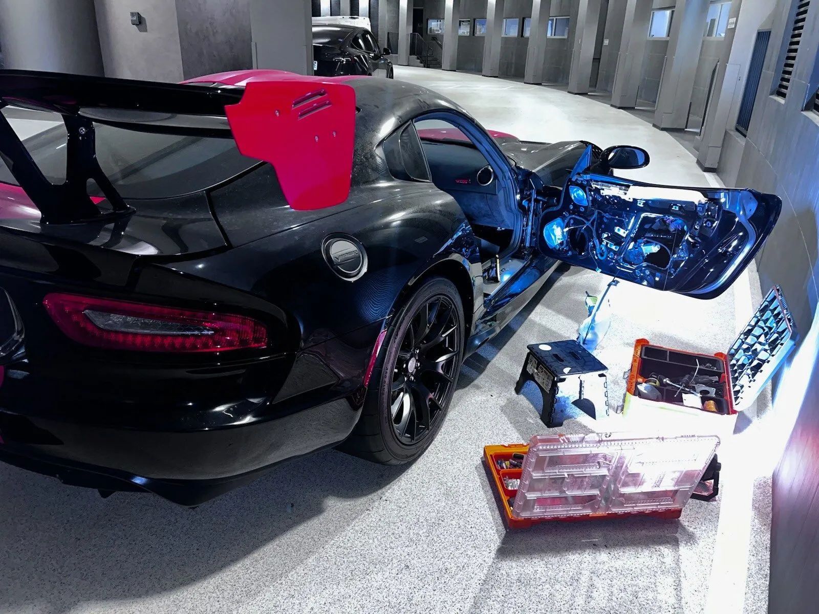 Black sports car with red accents, door open, surrounded by tools. Inside a showroom.