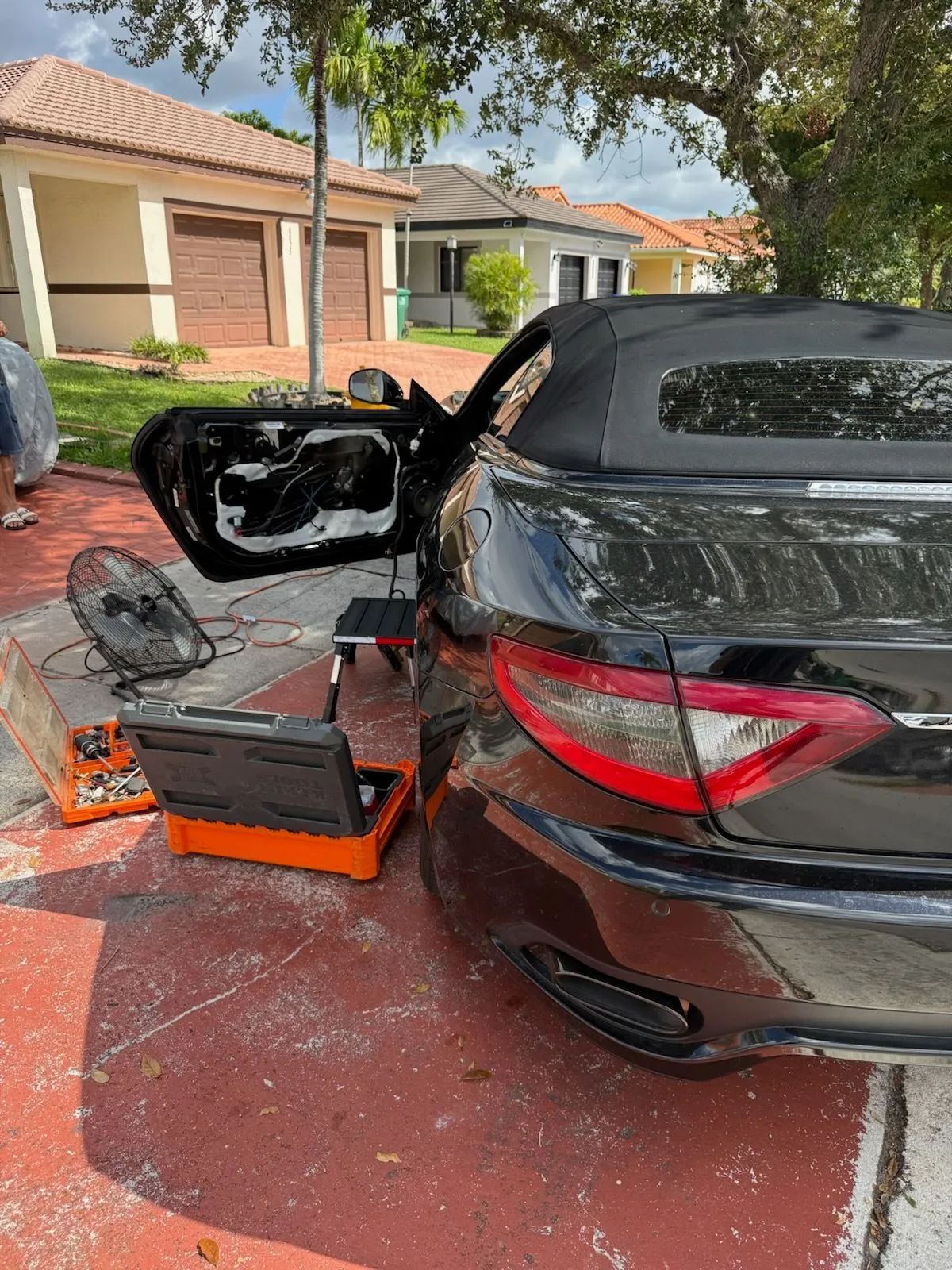 Black convertible car with door open, tools and parts visible in driveway.