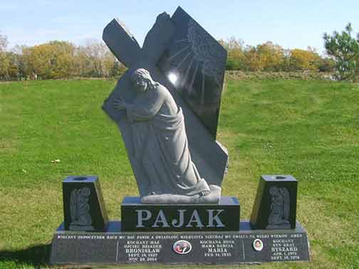 Monument with carved figure, cross, and names; black granite, grassy setting.
