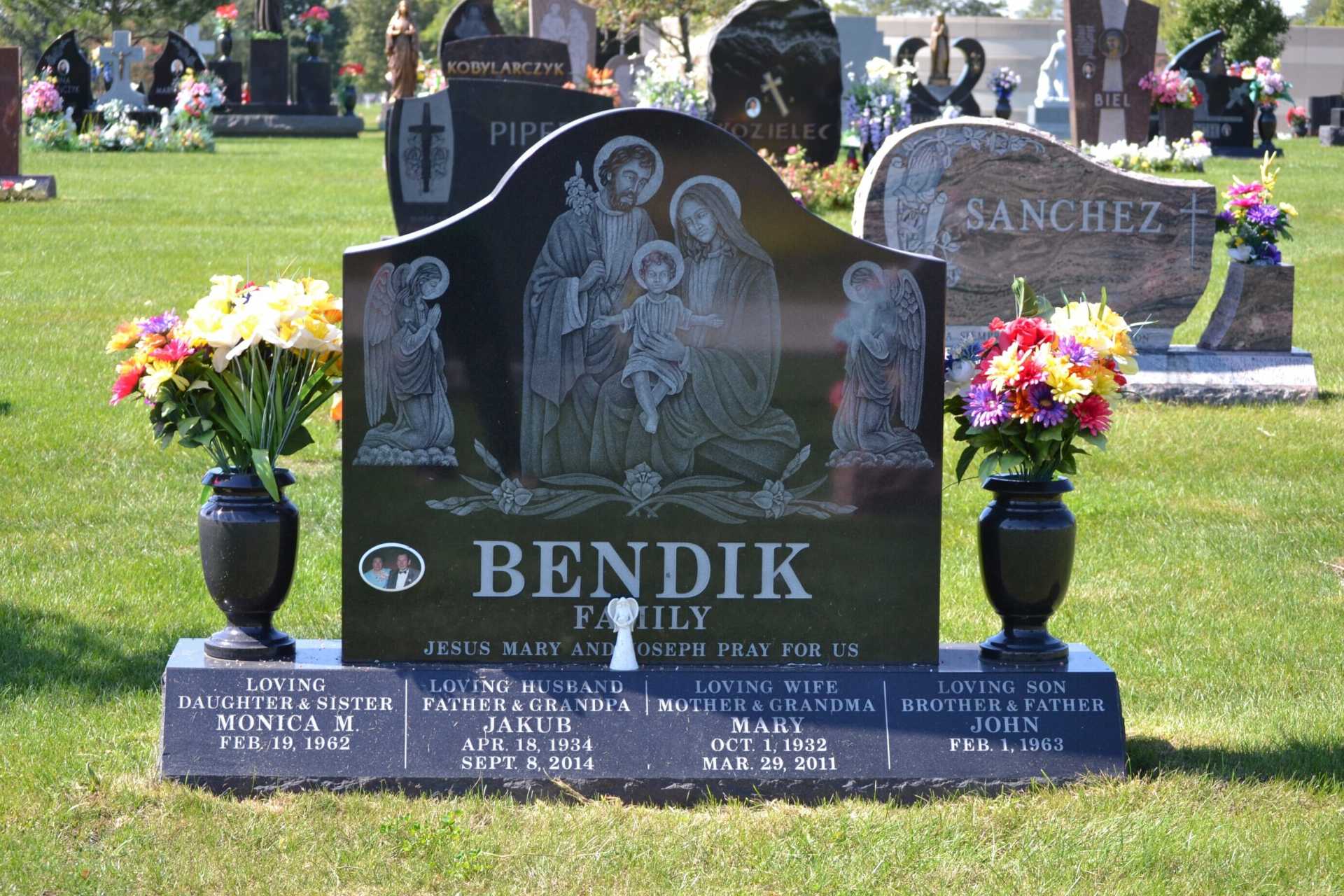 Black granite tombstone with religious family etching, flowers, and names in a cemetery.