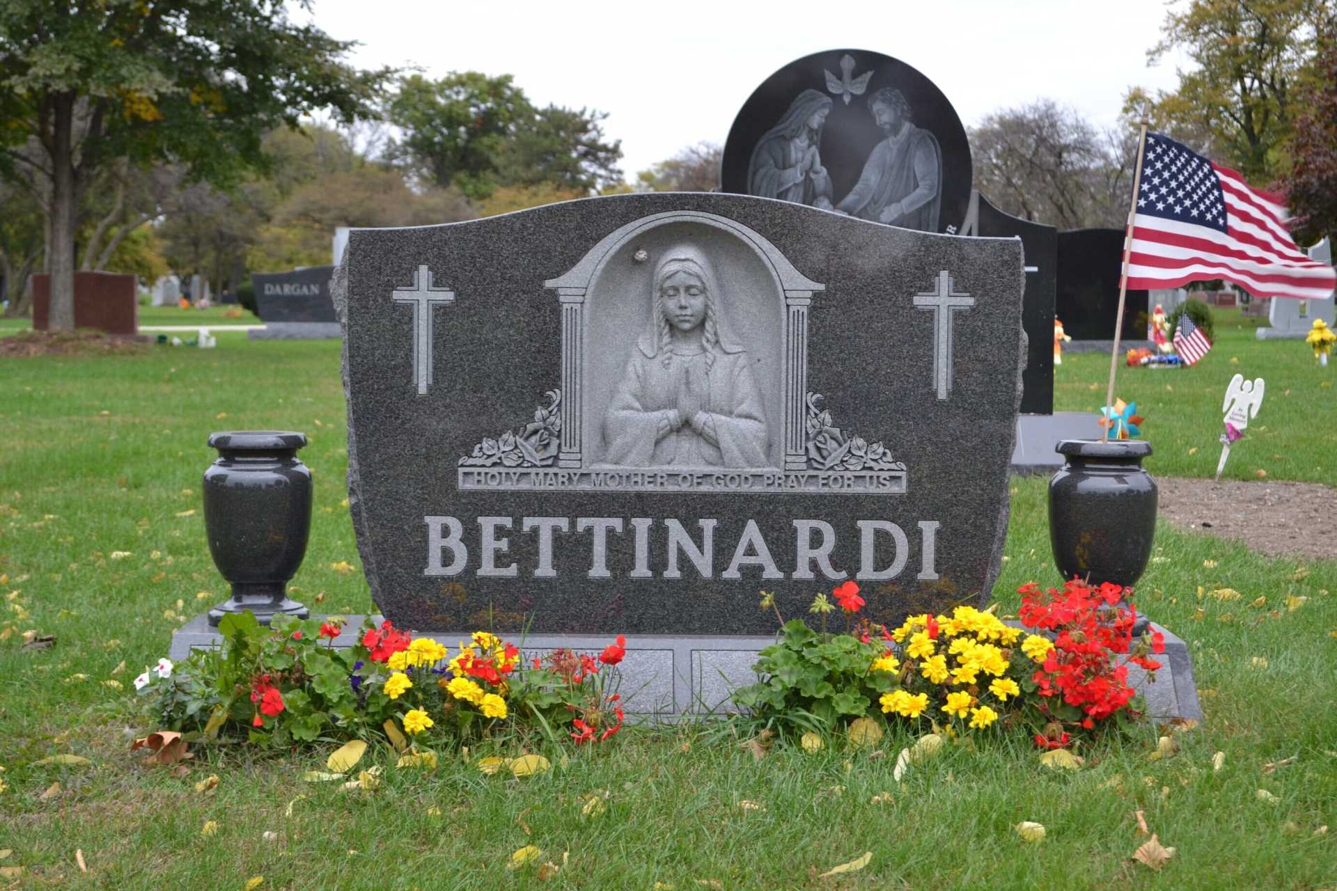 Tombstone of Bettinardi in a cemetery, with a sculpted portrait, cross, flowerbeds, and US flag.