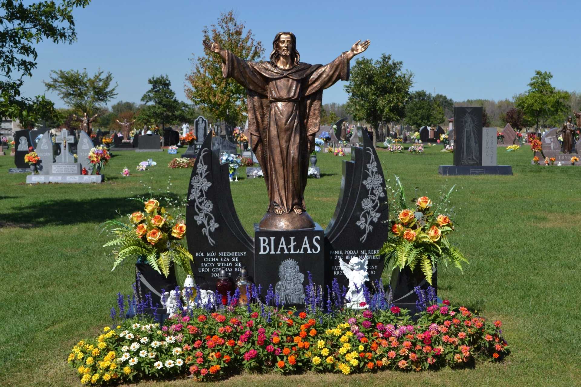 Bronze Jesus statue on ornate headstone in a cemetery, surrounded by flowers.