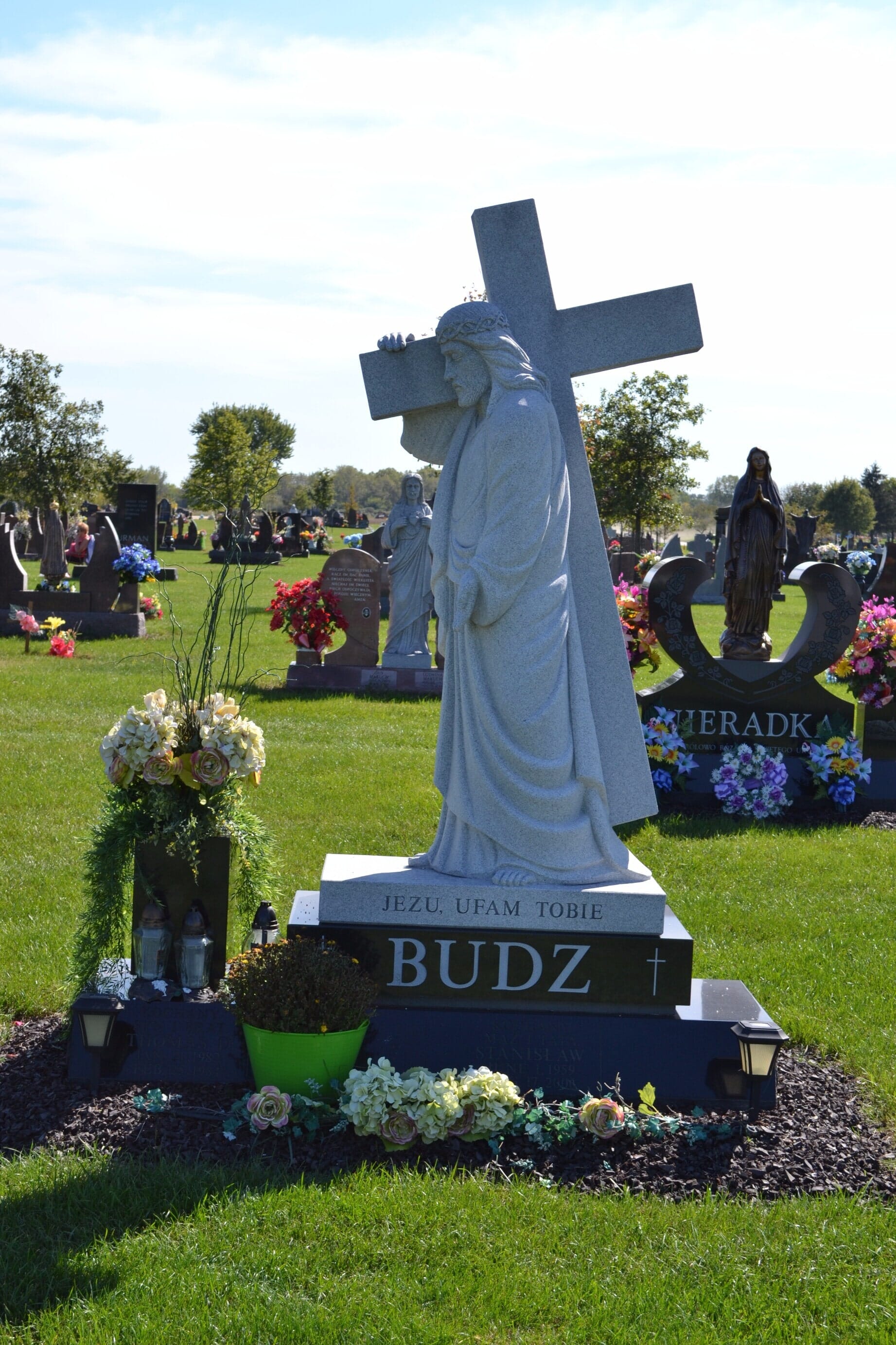 A granite grave marker featuring a statue of Jesus carrying a cross, surrounded by flowers in a cemetery.