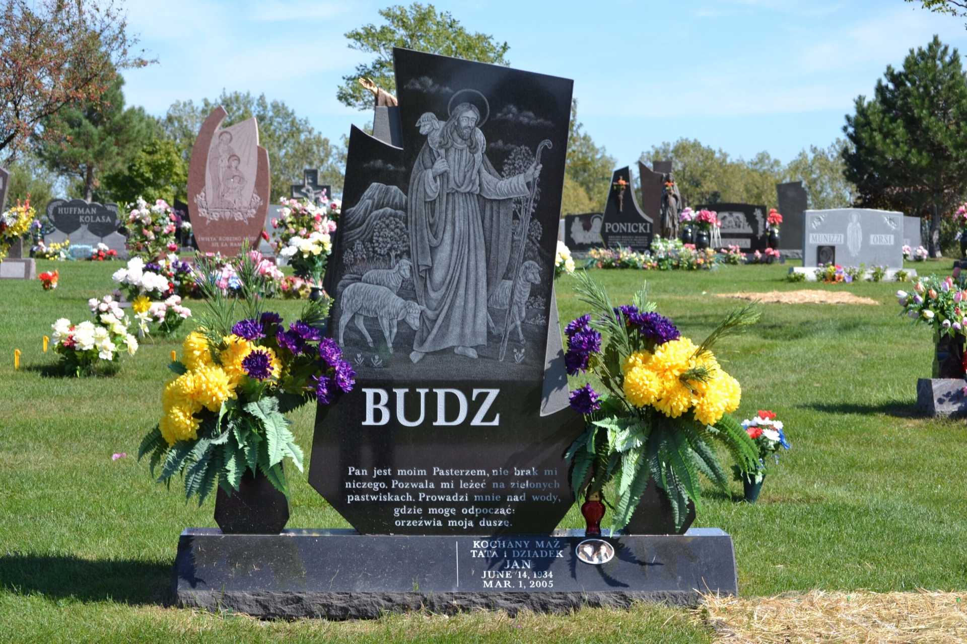 Black tombstone with the name BUDZ and an engraved image of a religious figure in a cemetery.