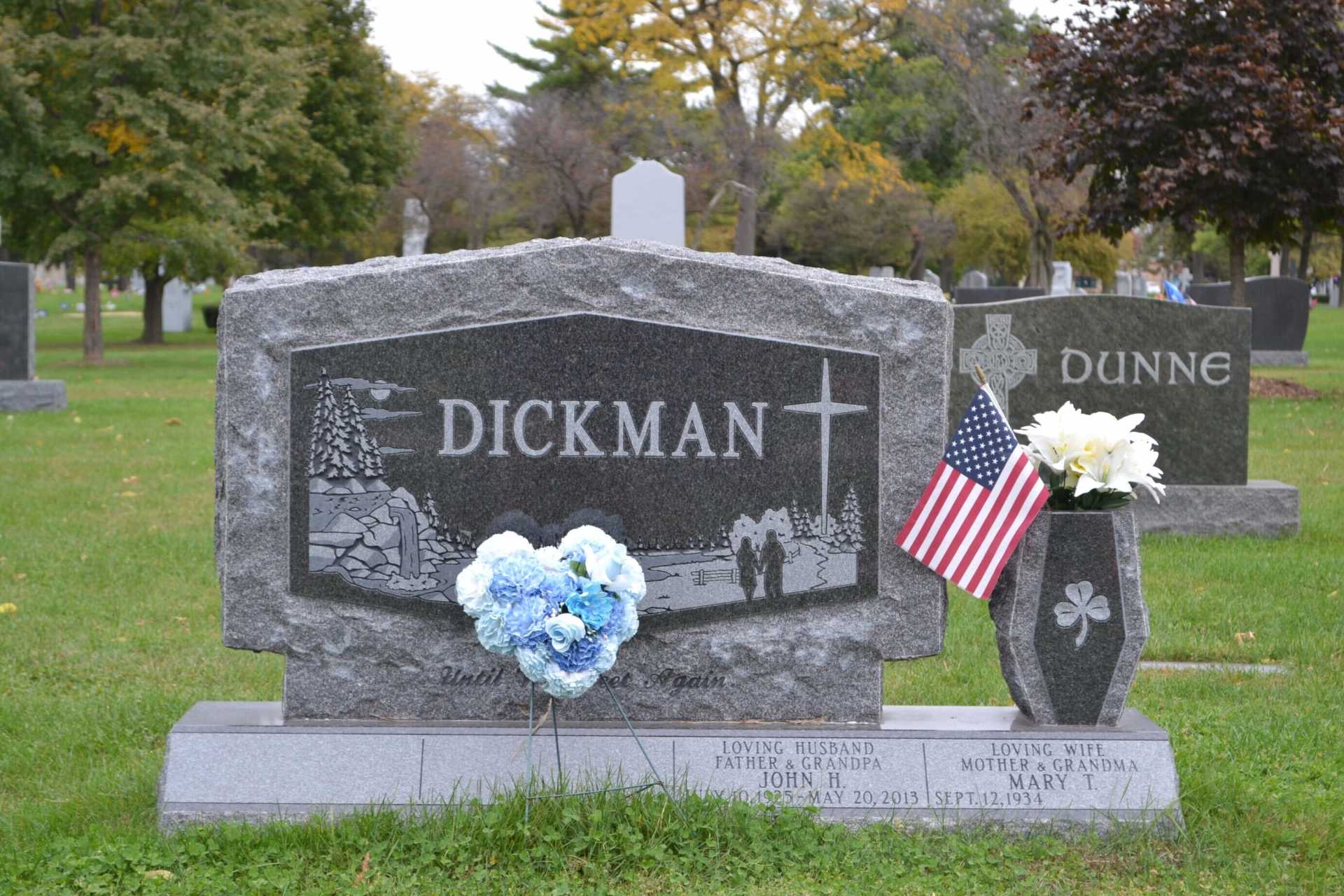 Grave marker for Dickman in a cemetery, with US flag and blue flower arrangement.
