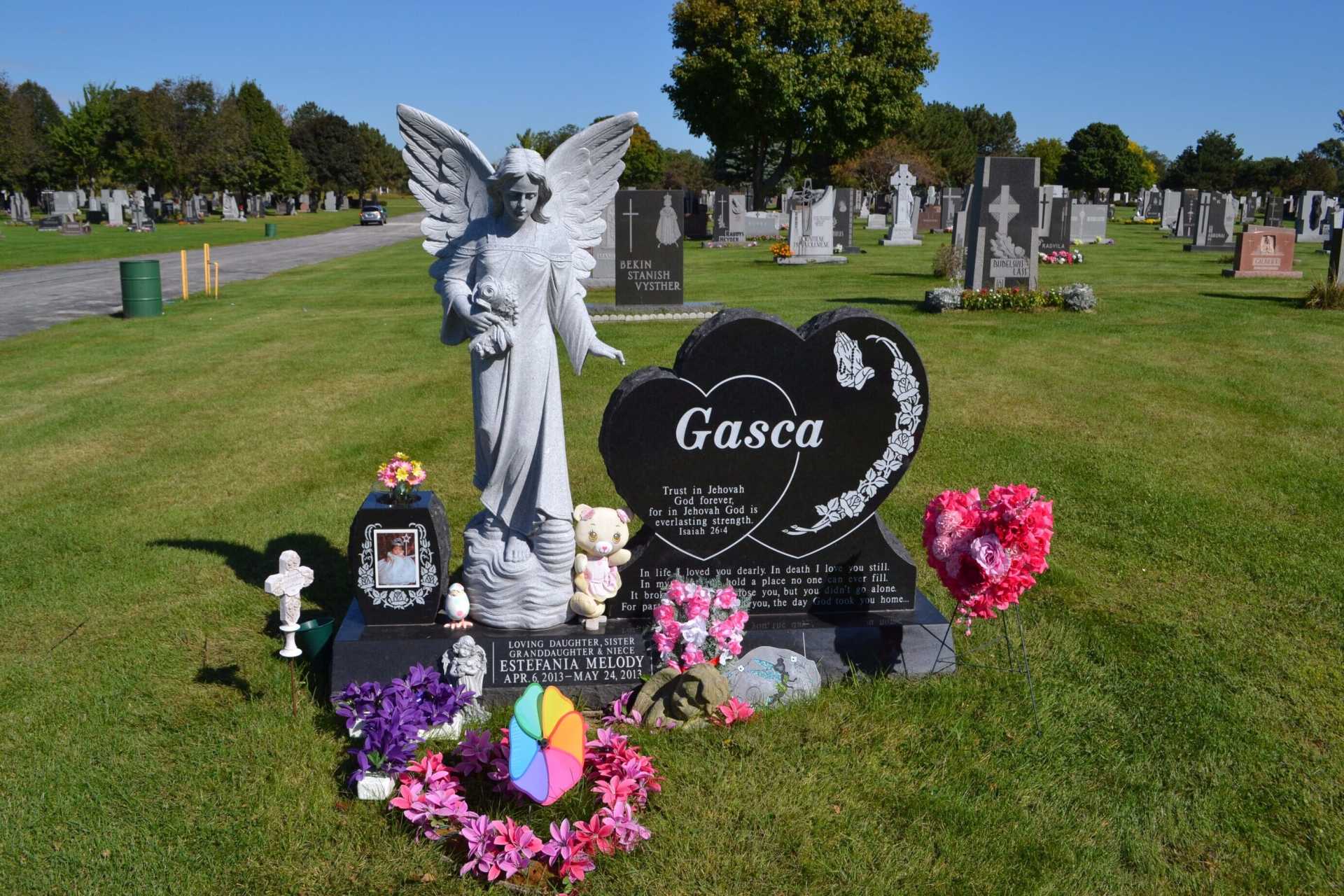 Grave with angel statue, heart-shaped headstone, and colorful flowers in a sunny cemetery.