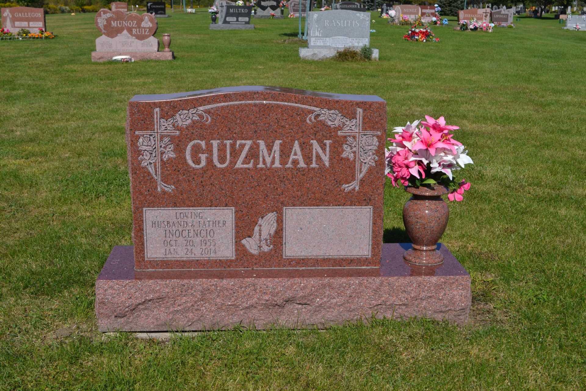Red Guzman headstone in a grassy cemetery with pink flowers.