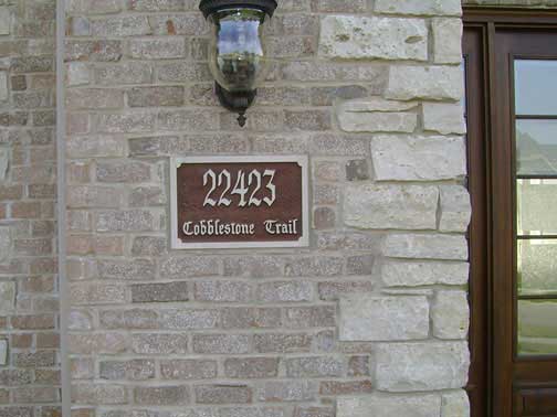 Address sign on brick and stone wall: 22423 Cobblestone Trail, brown numbers and text, under a light fixture, next to a door.