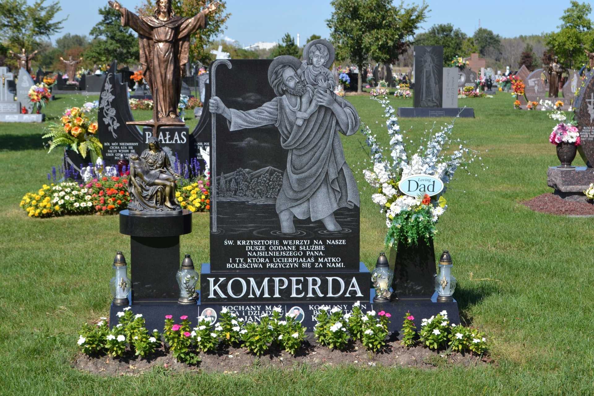 Gravestone with engraving of St. Christopher carrying a child, surrounded by flowers and candles in a sunny cemetery.