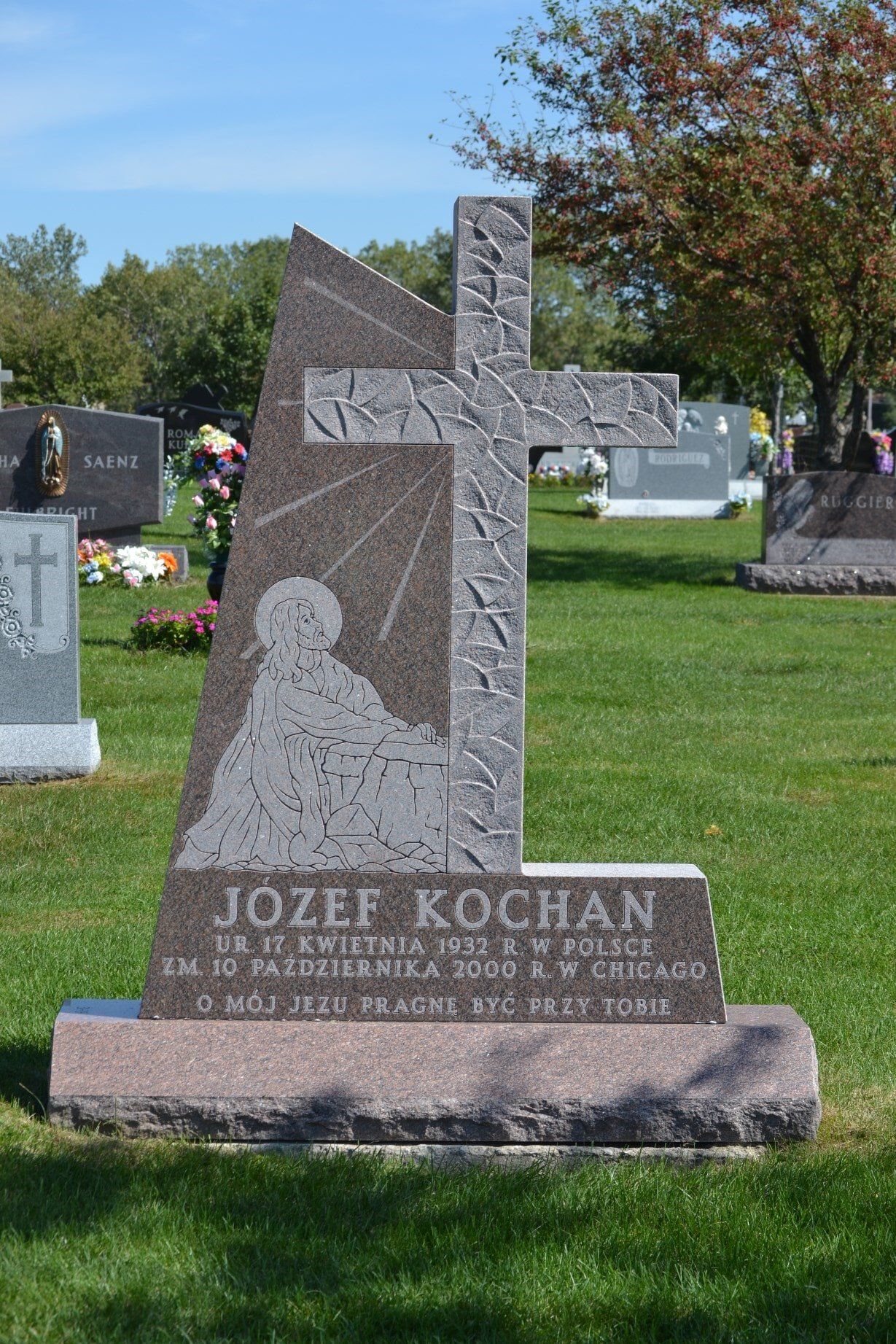 Grave of Jozef Kochan with cross and figure carving in a cemetery on a sunny day.