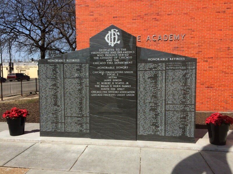 War memorial listing names, black stone, brick building in background, flowers in pots on the sides.