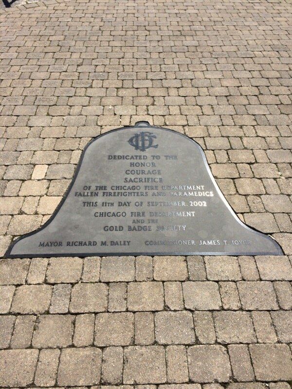 Memorial plaque for Chicago firefighters, shaped like a bell, set on brick pavers.