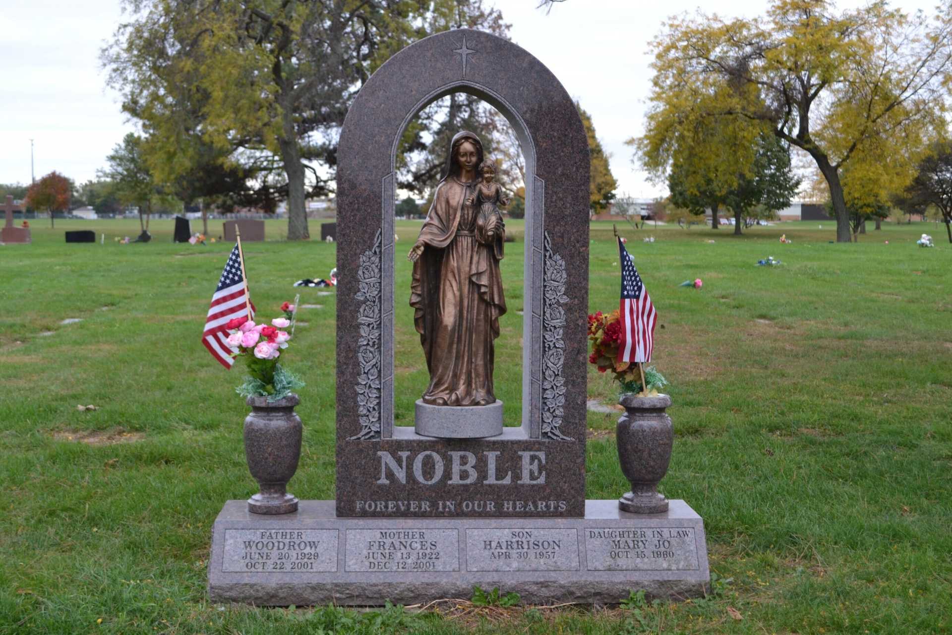 Tombstone with arched frame, bronze statue, and American flags in a grassy cemetery. Name on stone: Noble.