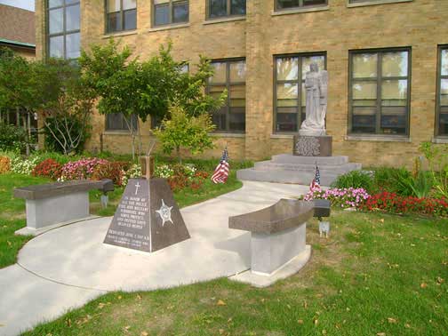 Courthouse grounds with a statue and memorial benches. Trees and flowers in the garden, flags and stone path.
