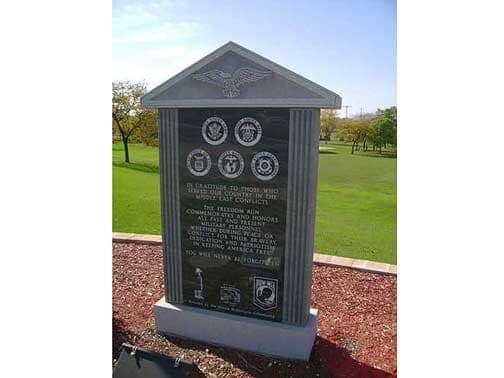 Monument with engraved text, military emblems, and a central eagle, set in a park with grass and trees.